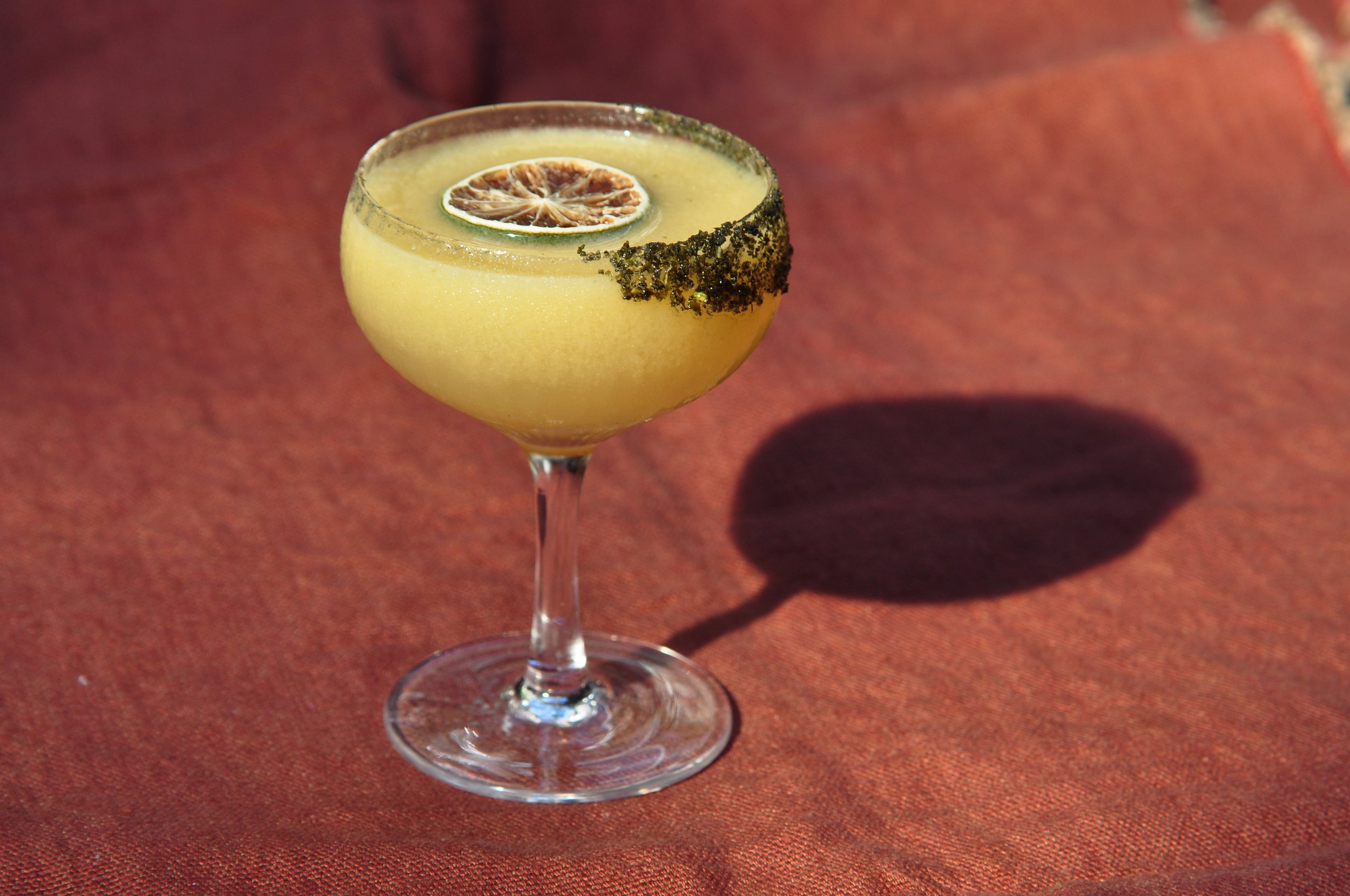 A yellow cocktail in a stemmed glass with a salted rim and a dried lime slice garnish on top, casting a shadow on a red fabric surface.