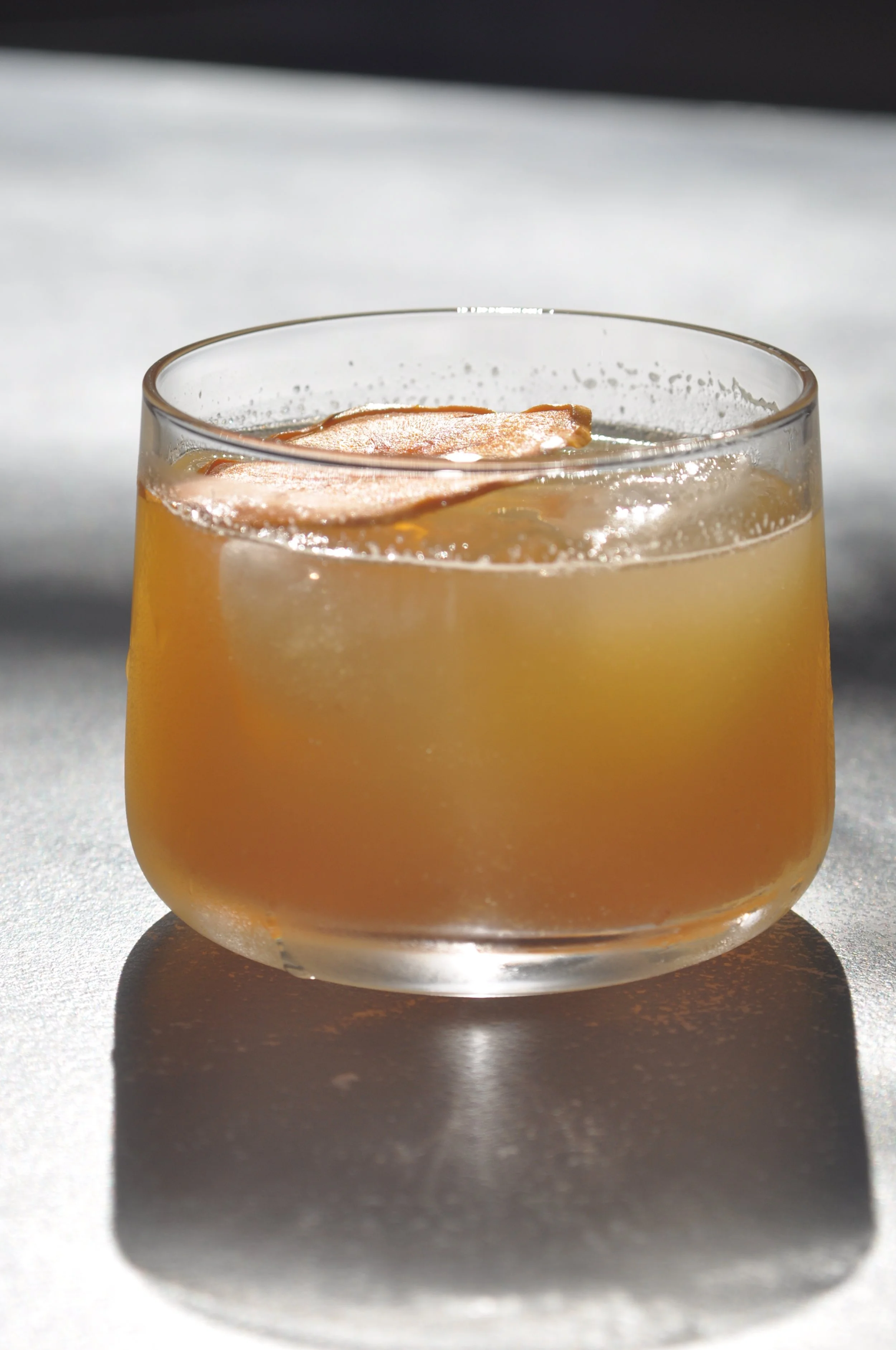 A glass of iced beverage with slices of apple on top, placed on a light surface with shadows and natural lighting.