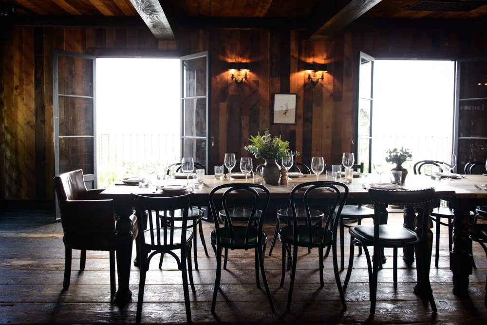 A rustic dining room with wooden walls and floor, featuring a long table set with glassware, plates, and flower arrangements, illuminated by natural light from open windows.