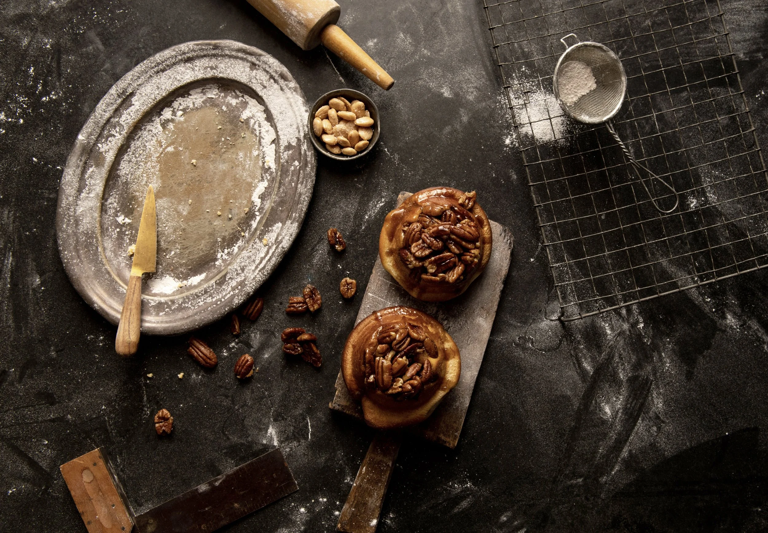 Two baked apples topped with pecans, placed on a baking tray on a dark, flour-dusted surface, with baking tools such as a rolling pin, knife, sifter, and cooling rack scattered around.