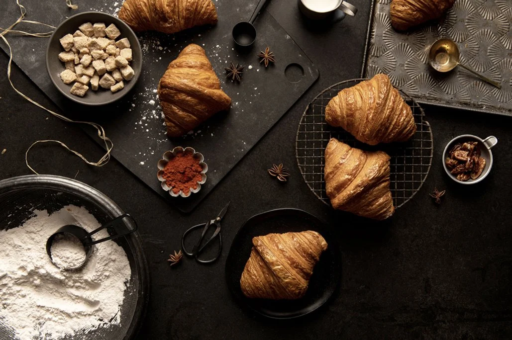 Fresh croissants on a wire rack and plate surrounded by baking ingredients like flour, sugar cubes, spices, and baking tools on a dark countertop.