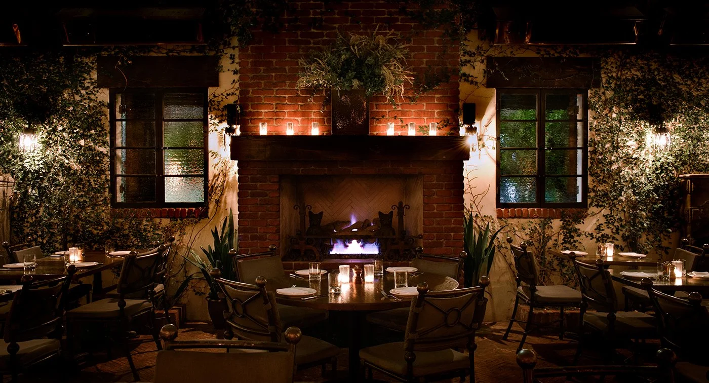 Interior of a cozy restaurant with a brick fireplace, surrounded by tables set with candles, and decorated with greenery and candles on the mantle.