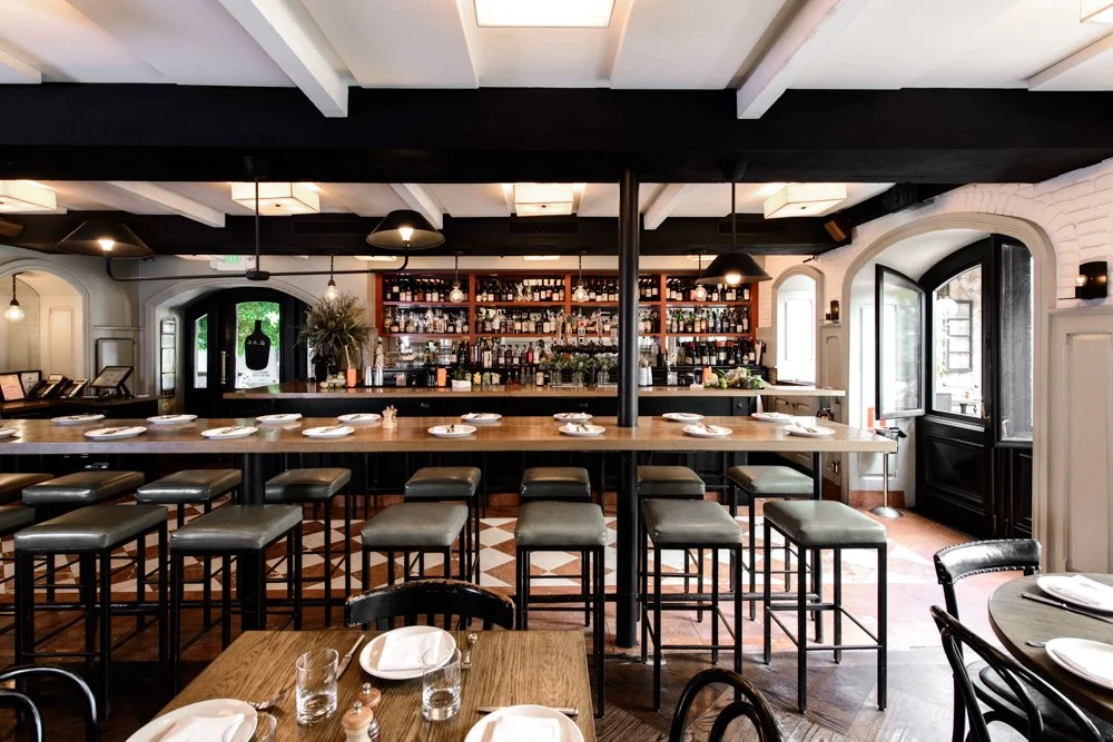 Empty restaurant with a bar, high stools, and tables set with plates and utensils.