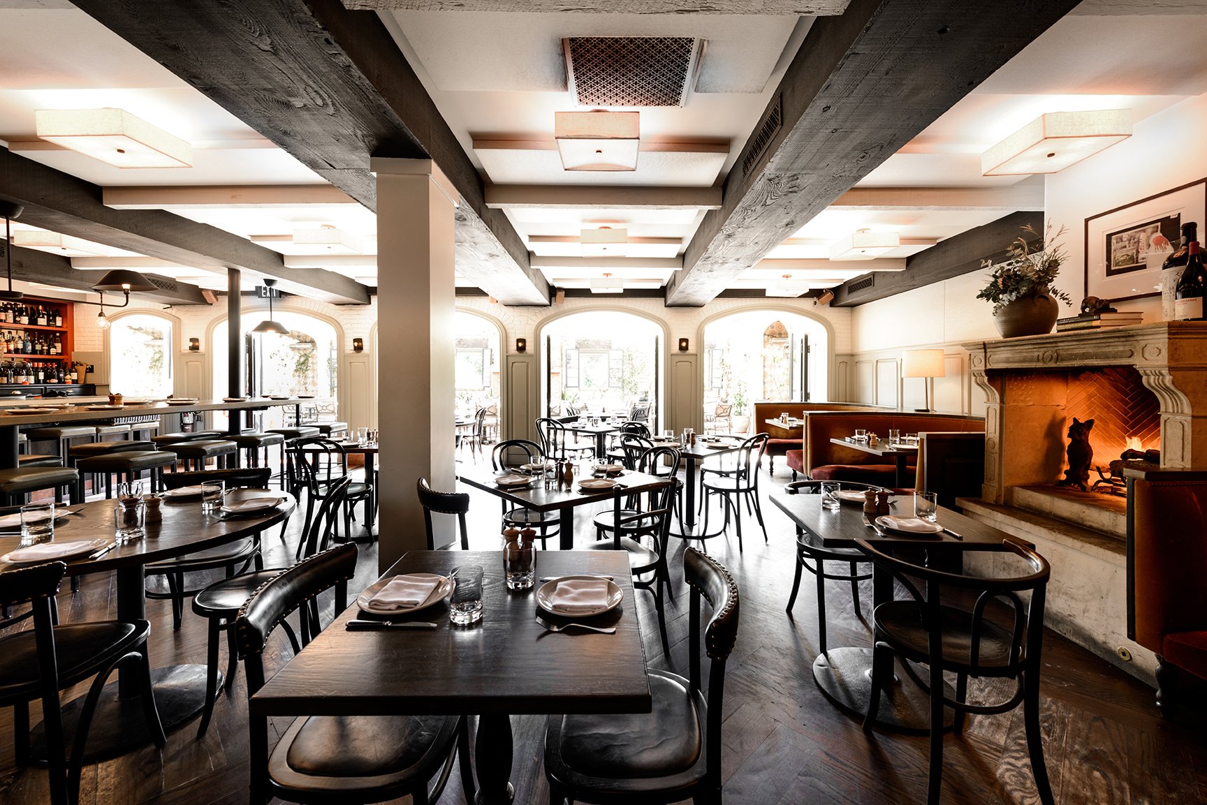 Elegant restaurant interior with wooden tables and black chairs, large windows providing natural light, a fireplace with decorations, and a bar area in the background.