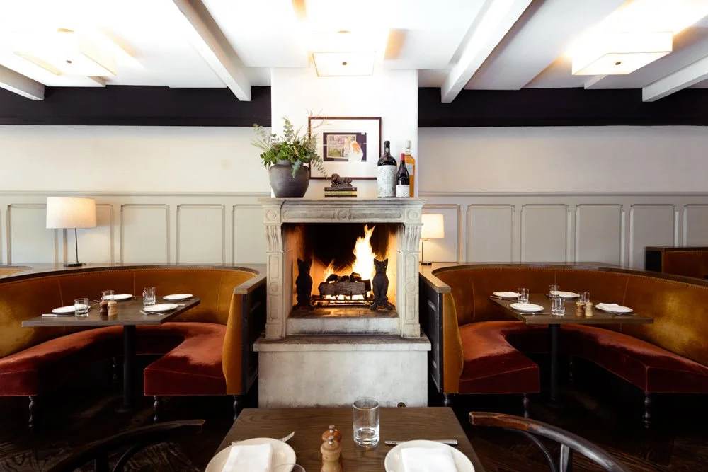 A cozy restaurant interior with a central fireplace, surrounded by curved booth seating, tables set with plates, glasses, and cutlery, and soft lighting from lamps and ceiling fixtures.