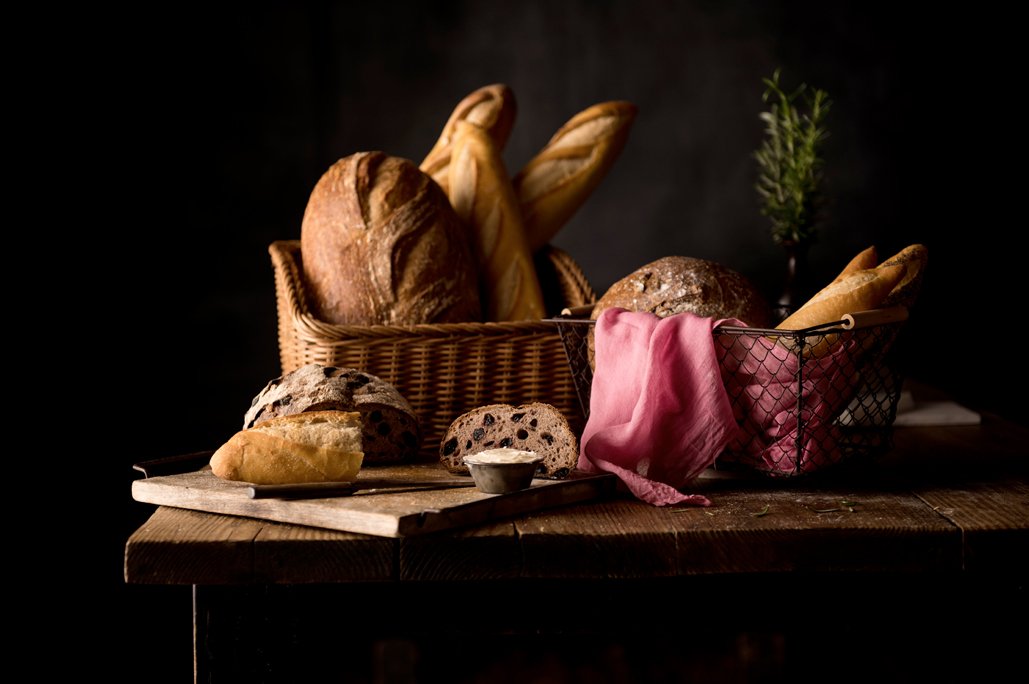 A variety of bread types including loaves and rolls in woven baskets and on a wooden table, with a small cup of butter and a sprig of greenery for decor.