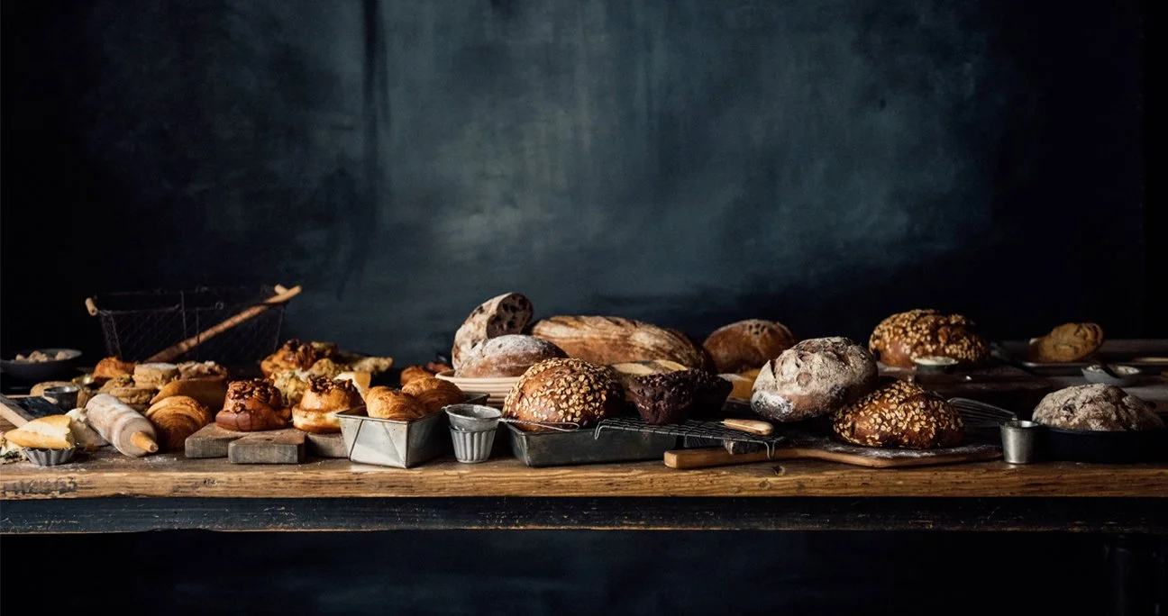 Various types of bread and baked goods spread across a rustic wooden table against a dark background.