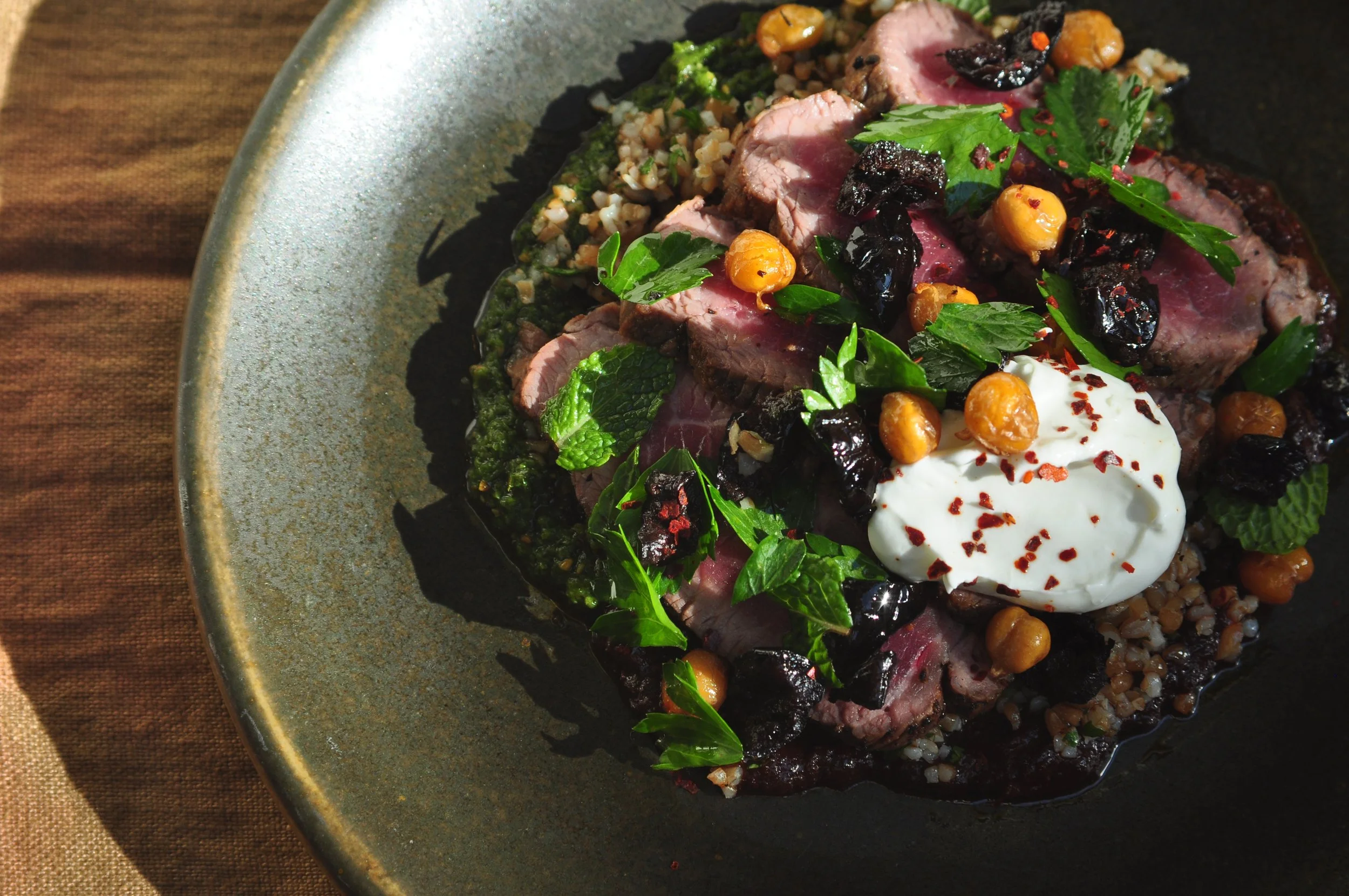 Plate of sliced seared beef with herbs, chickpeas, black olives, a dollop of white sauce garnished with red pepper flakes, and a bed of grains.
