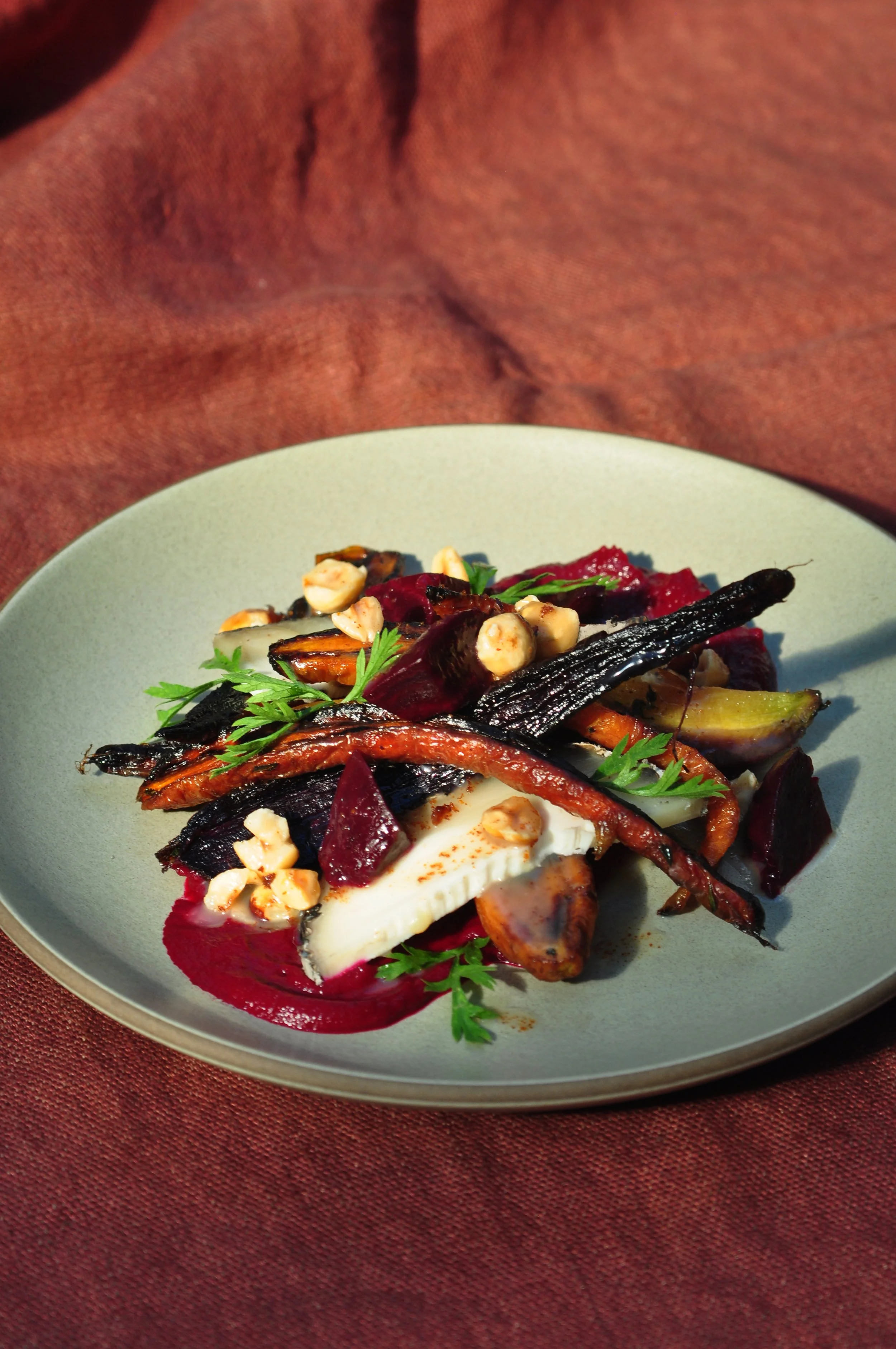 A plate of roasted vegetables, goat cheese, garnished with herbs and nuts, on a gray plate with a reddish fabric background.