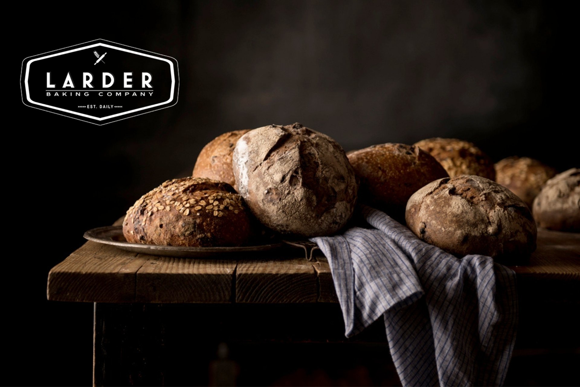 A variety of rustic bread loaves on a wooden table with a dark background, some topped with oats, and a striped cloth underneath.
