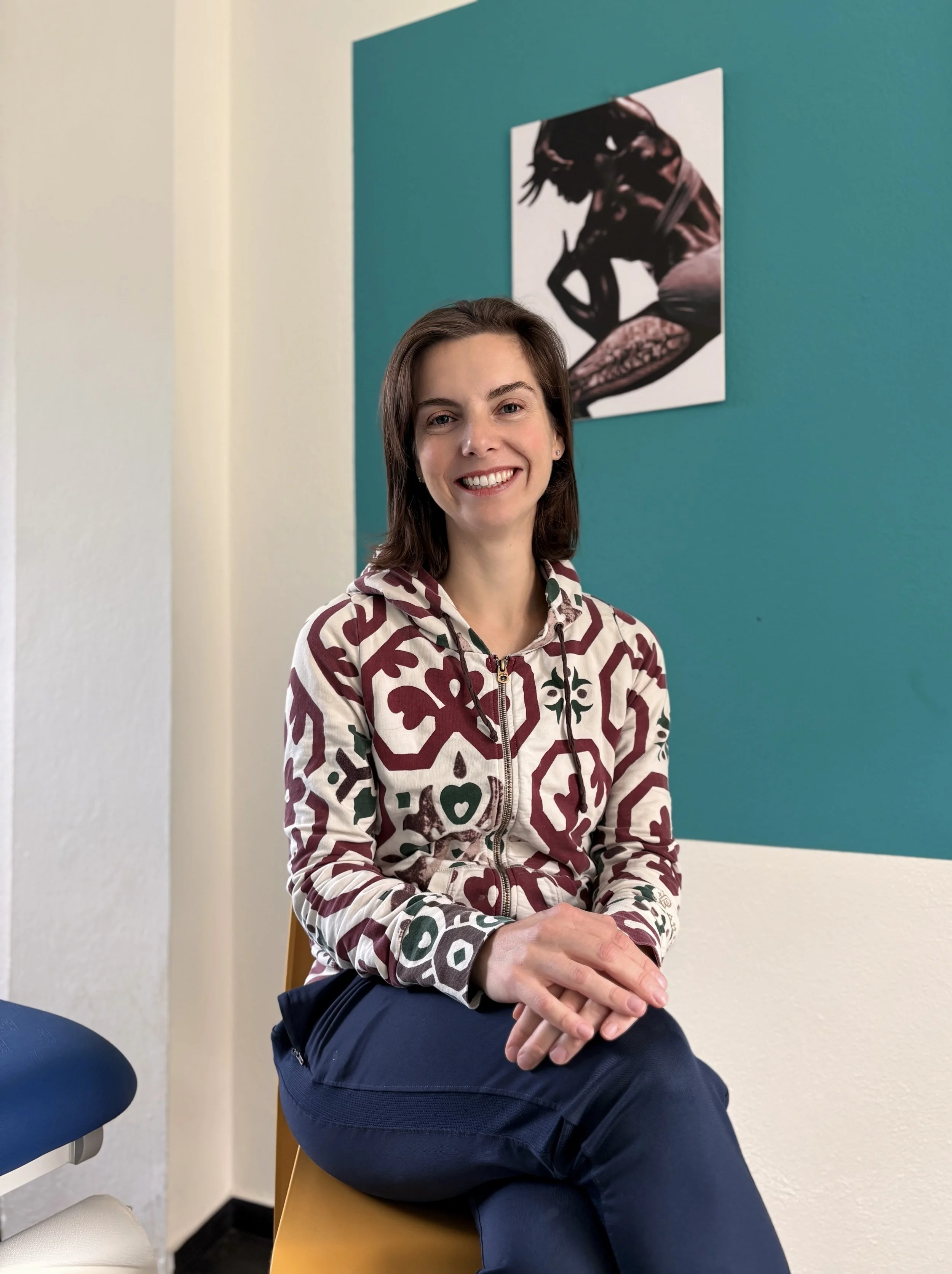 Una donna sorridente seduta su una sedia di legno in un ambiente con pareti bianche e una parete azzurra, con un quadro astratto appeso.
