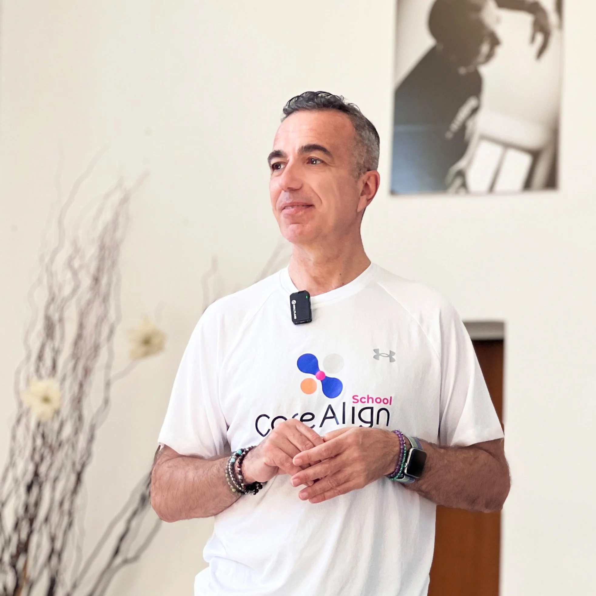 Uomo con capelli corti e barba, indossa una maglietta bianca con il logo di School coAlign, braccia incrociate, indossa bracciali e un orologio, sfondo chiaro con poster nero e bianco.
