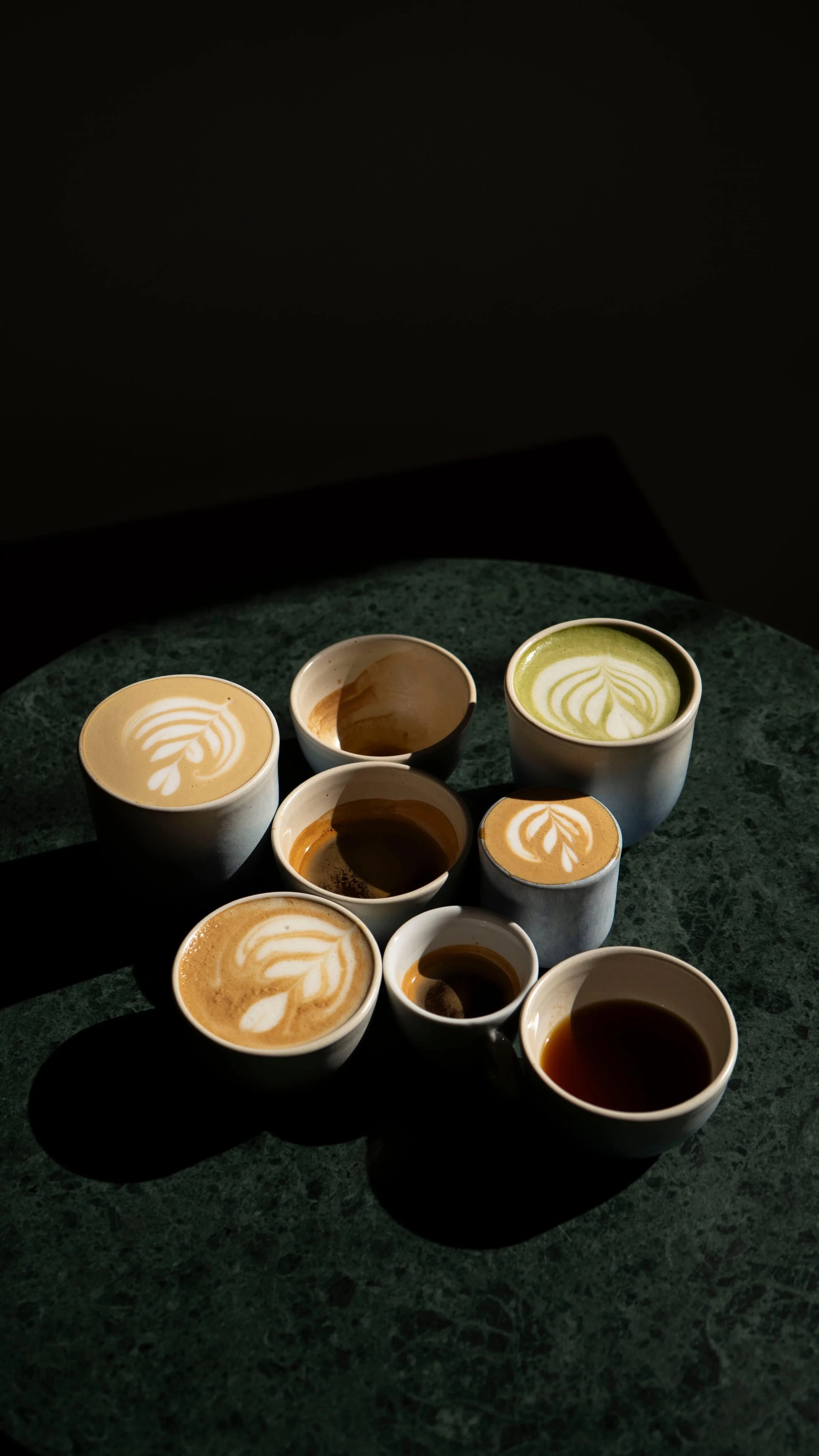 Several cups of coffee with different latte art designs on top, placed on a dark green marble table in a dimly lit setting.