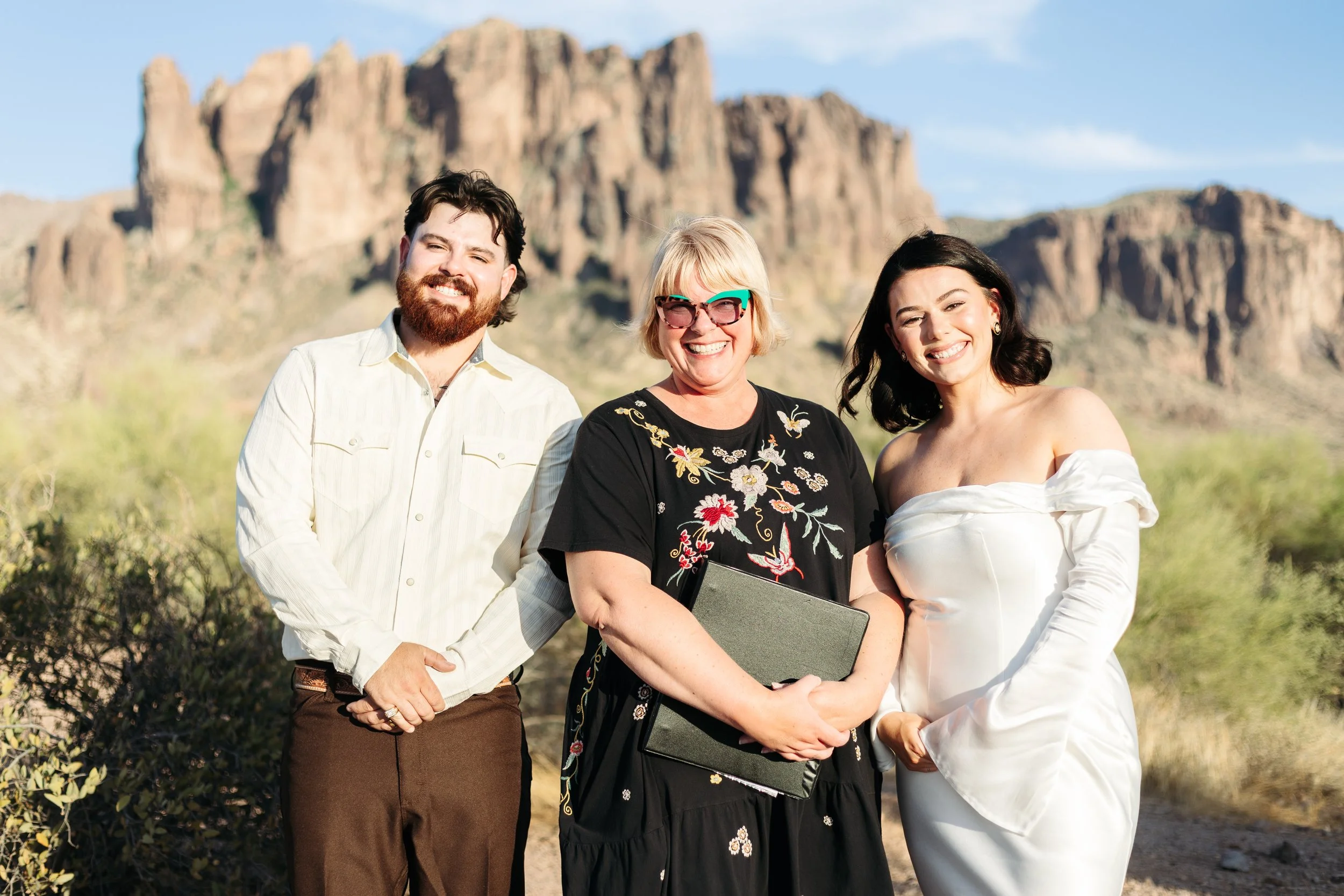 Three people standing outdoors with desert landscape and mountains in the background, smiling at the camera.
