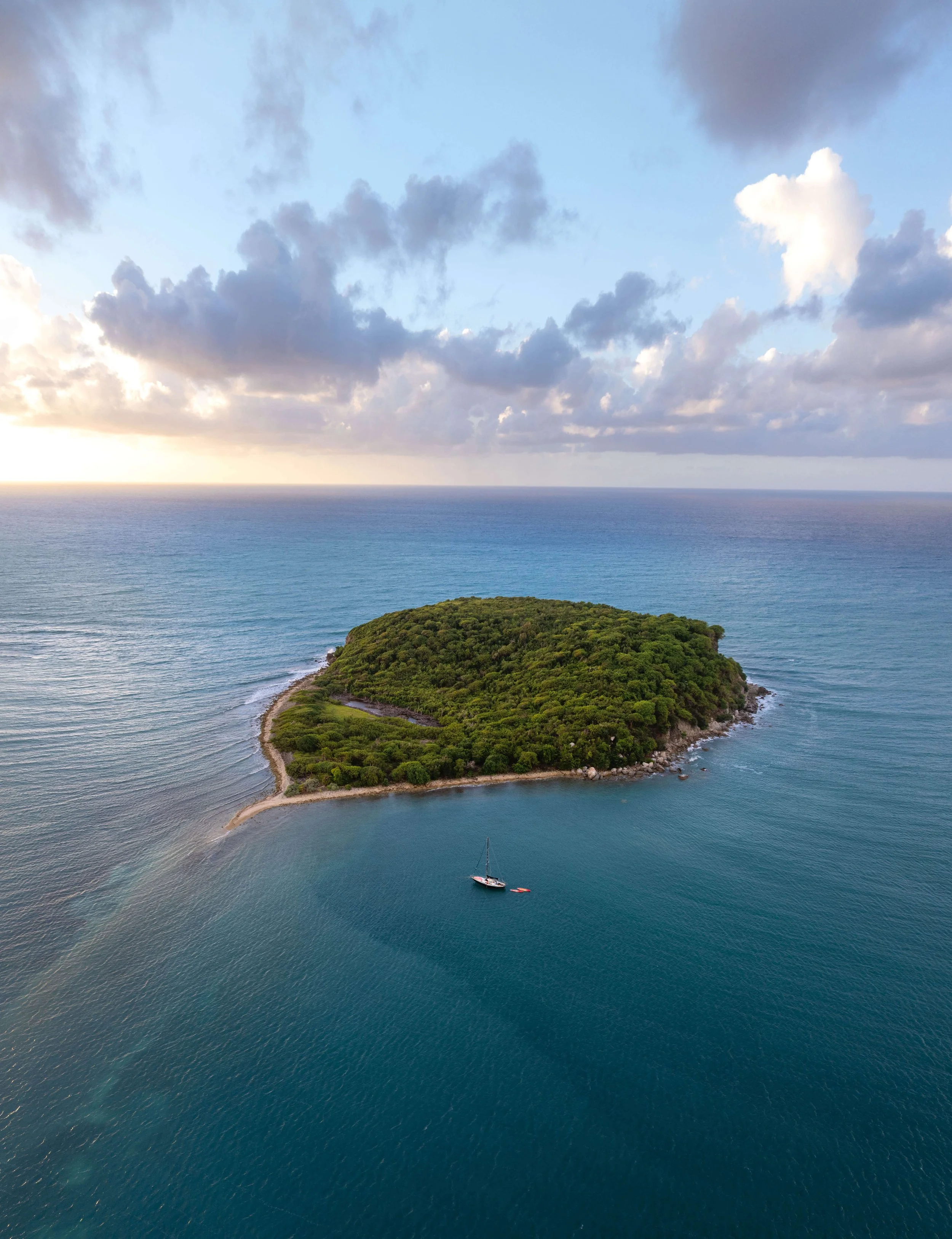 Aerial view of a small lush green island in the ocean with a sailboat nearby, under a partly cloudy sky.