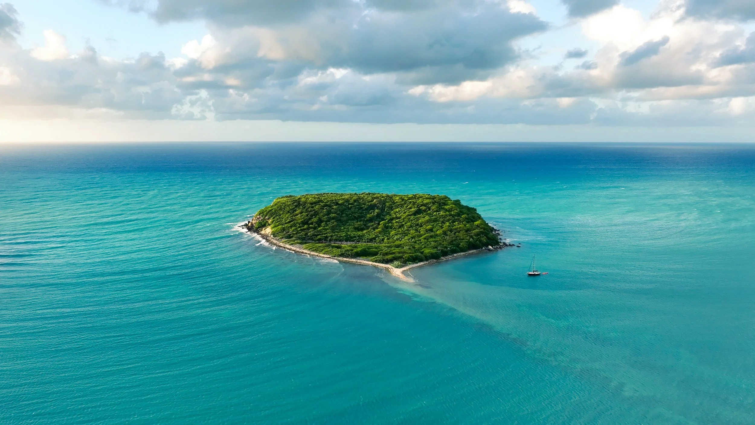 A small, green island surrounded by blue ocean water with a sailboat near the island and cloudy sky above.