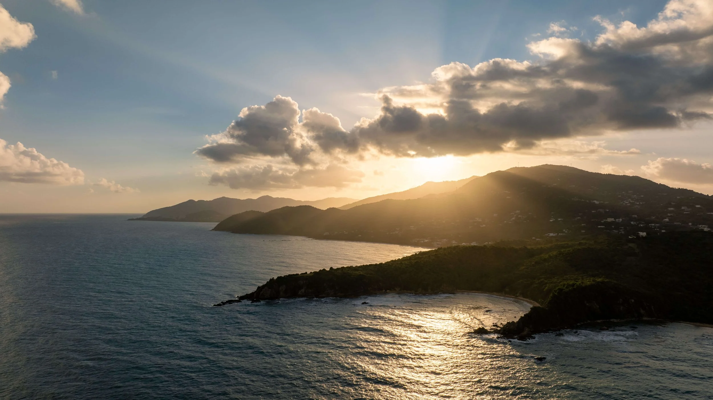 Sunset over the ocean with hills and mountains along the coast, partly cloudy sky, and sunlight reflecting on the water.