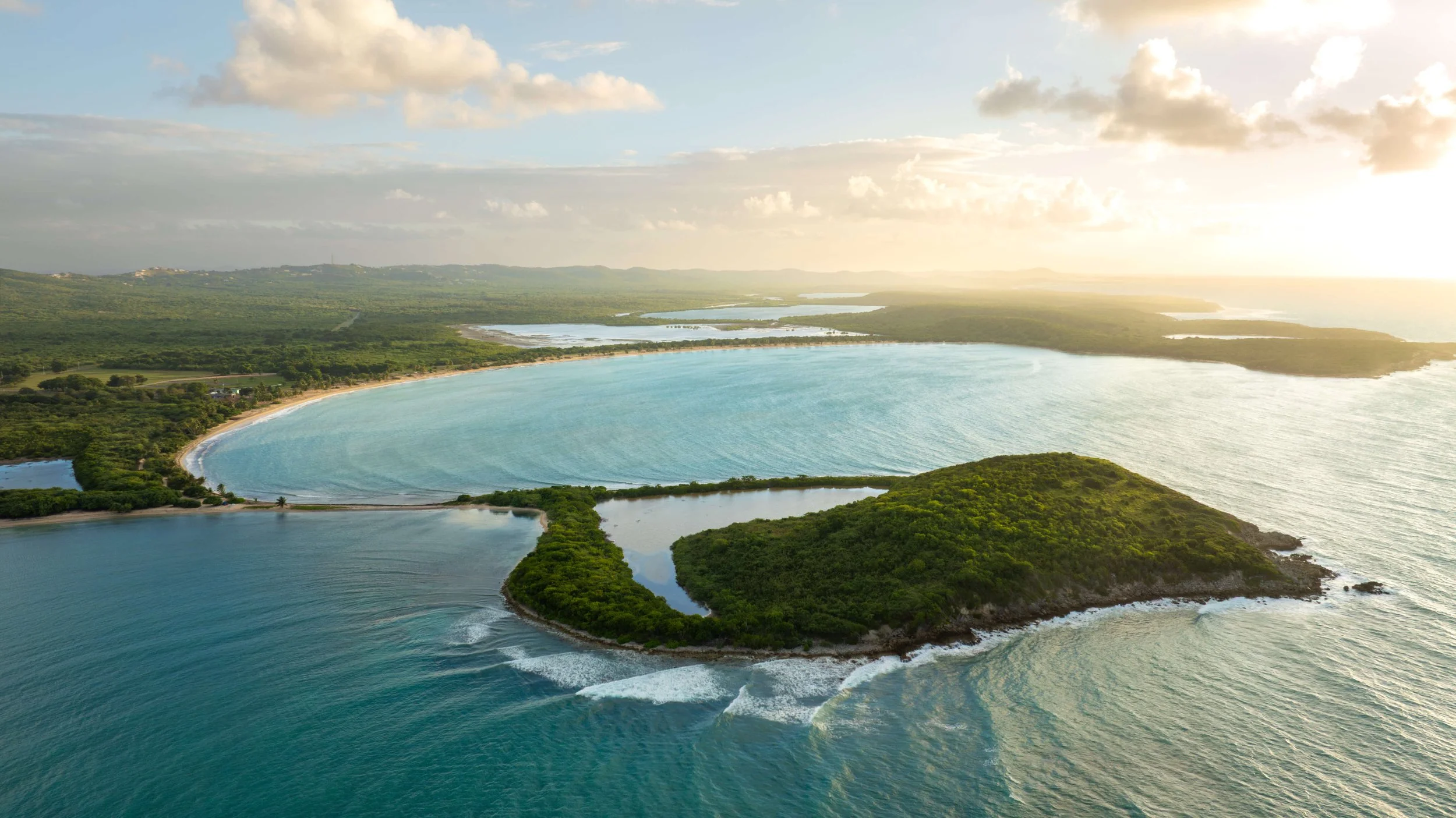 An aerial view of a coastal landscape featuring a large crescent-shaped beach, a body of water with lush green islands, and a scenic horizon with clouds and sunlight.