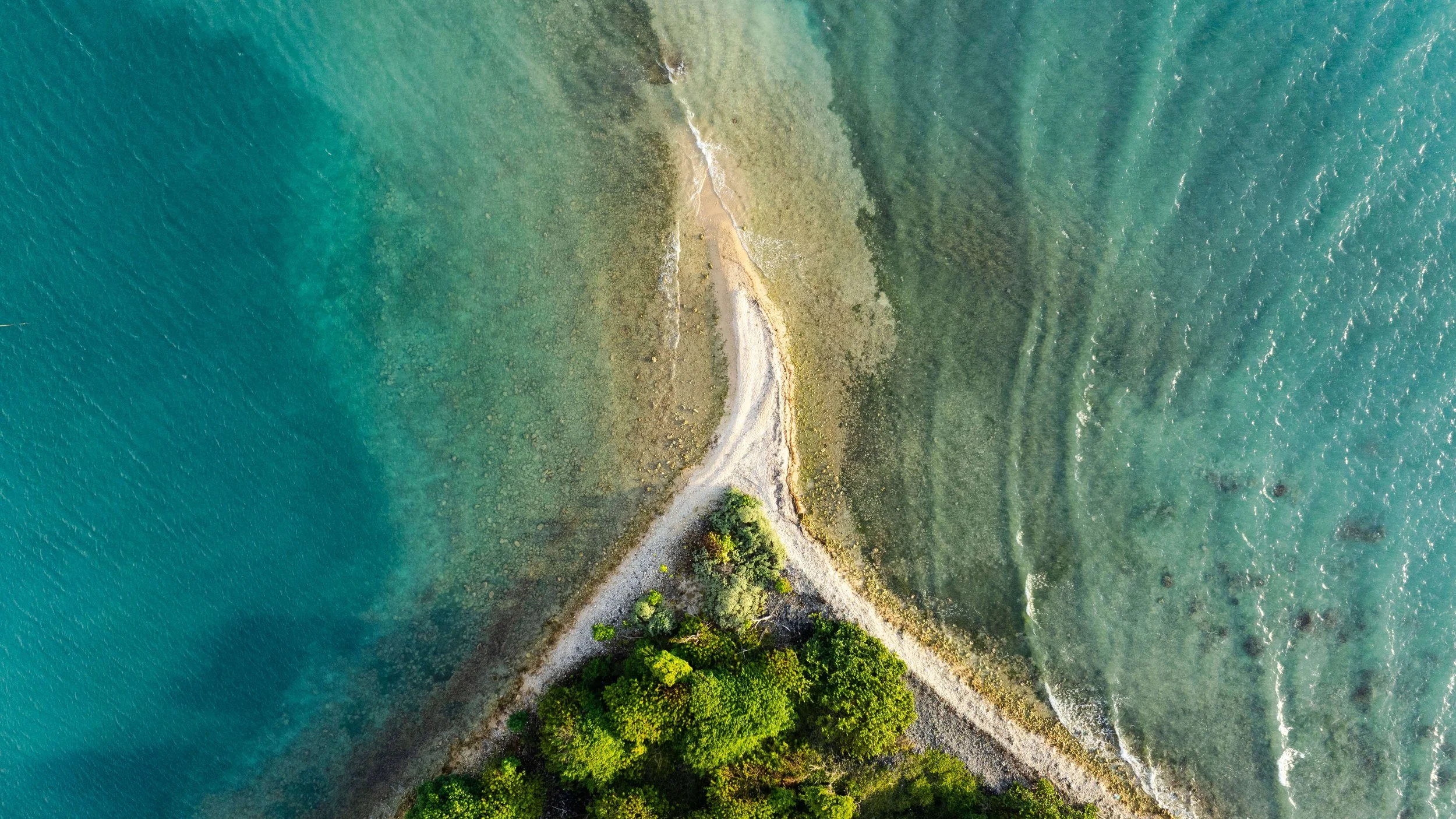 Aerial view of a coastal scene showing a narrow peninsula with trees, a gravel path, and a shoreline meeting the ocean with clear blue-green water.