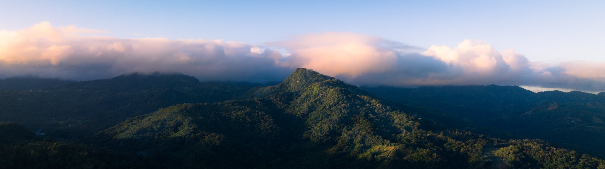 Aerial photo of a sunrise in the green mountains of central Puerto Rico. The top third of the image is sky with tones of blue and orange/pink tone clouds. There are part of the mountains still in the shadows because of the low angle of the sun