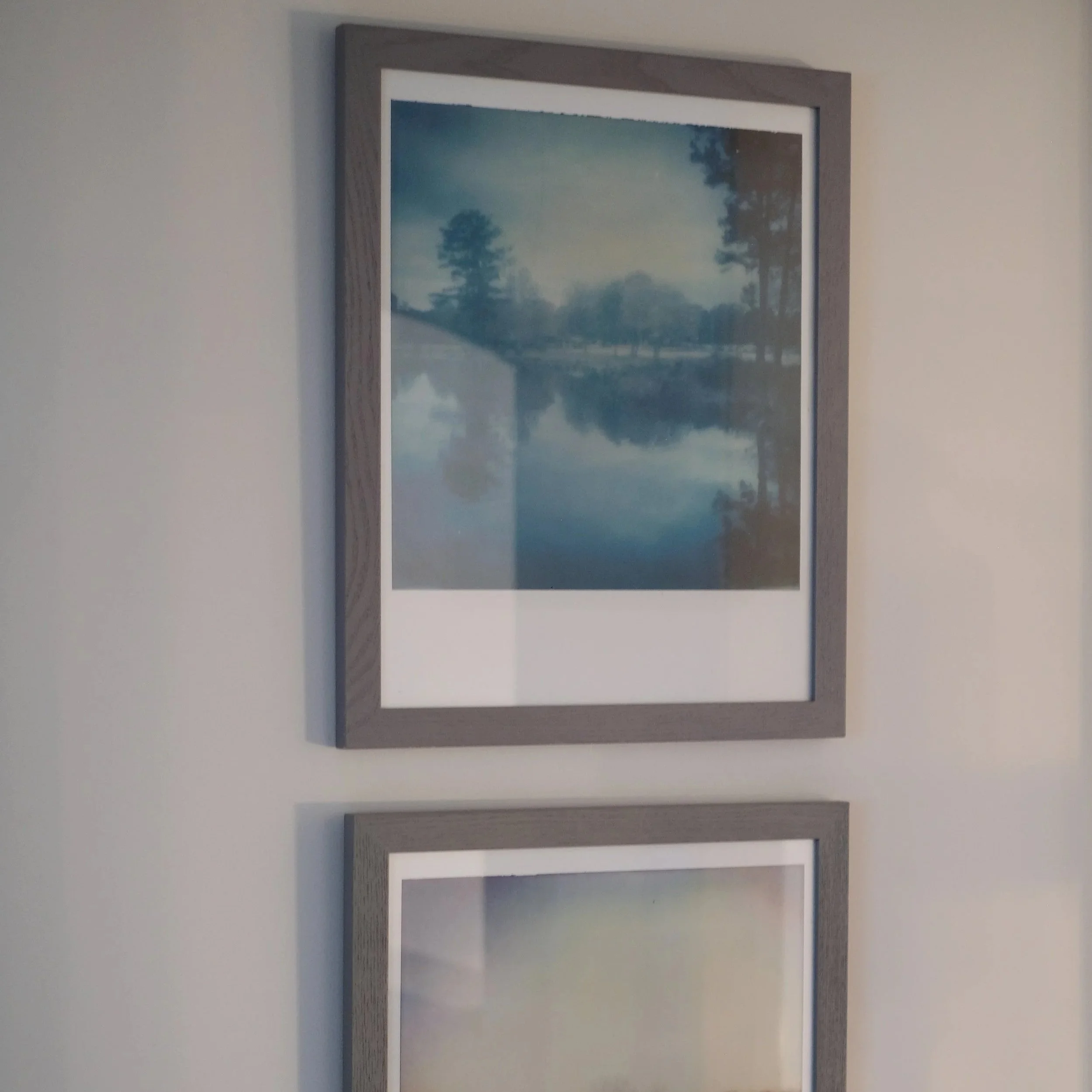 Framed landscape photo of a lake with trees and cloudy sky reflected on the water, mounted on a light-colored wall.