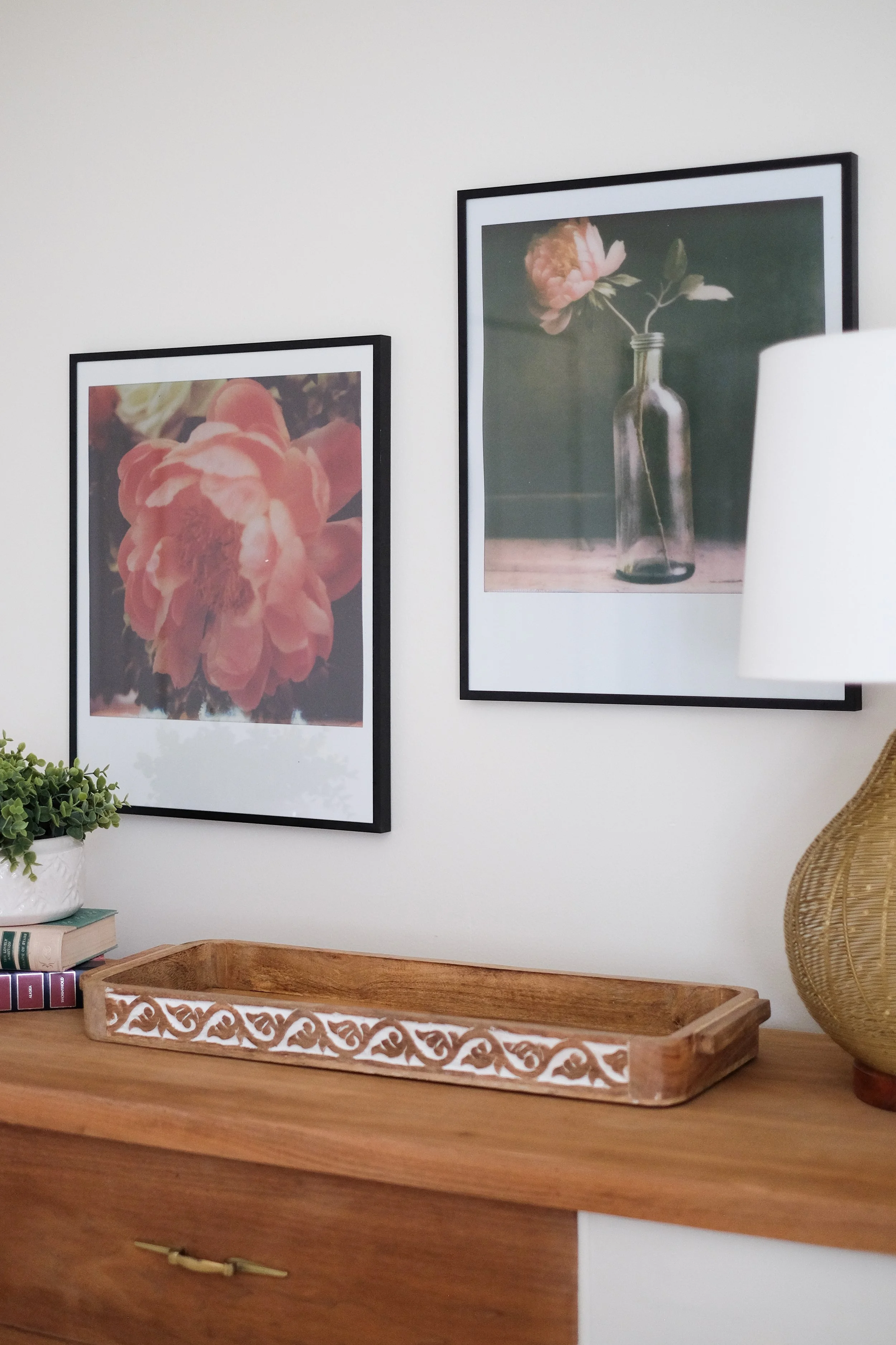 Decorative interior with two framed floral photographs on a white wall, a wooden sideboard, a potted plant, a stack of books, and a tall woven lamp.