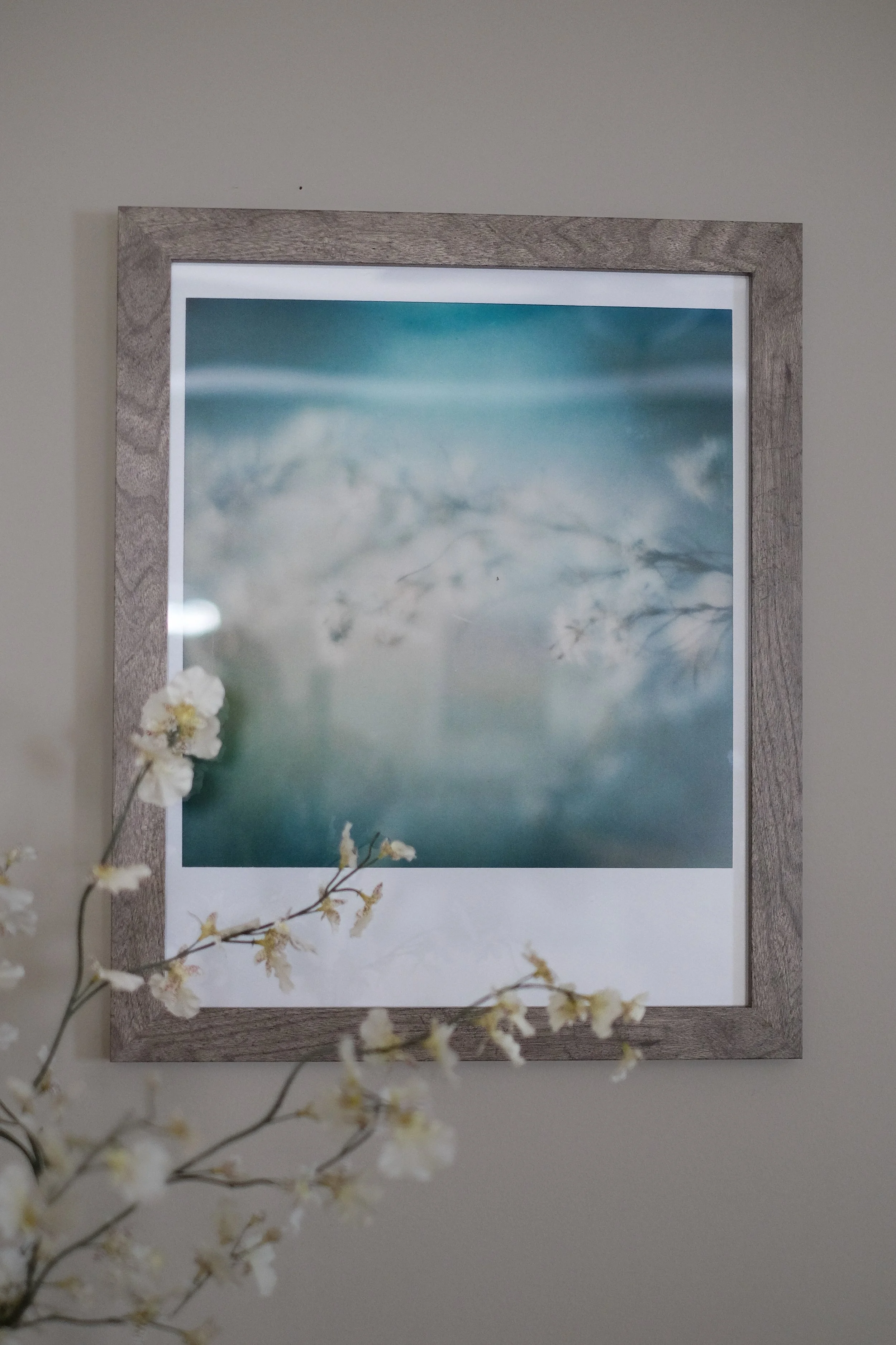 Framed abstract artwork with soft blue and white colors, partially obscured by a branch with white flowers in the foreground.