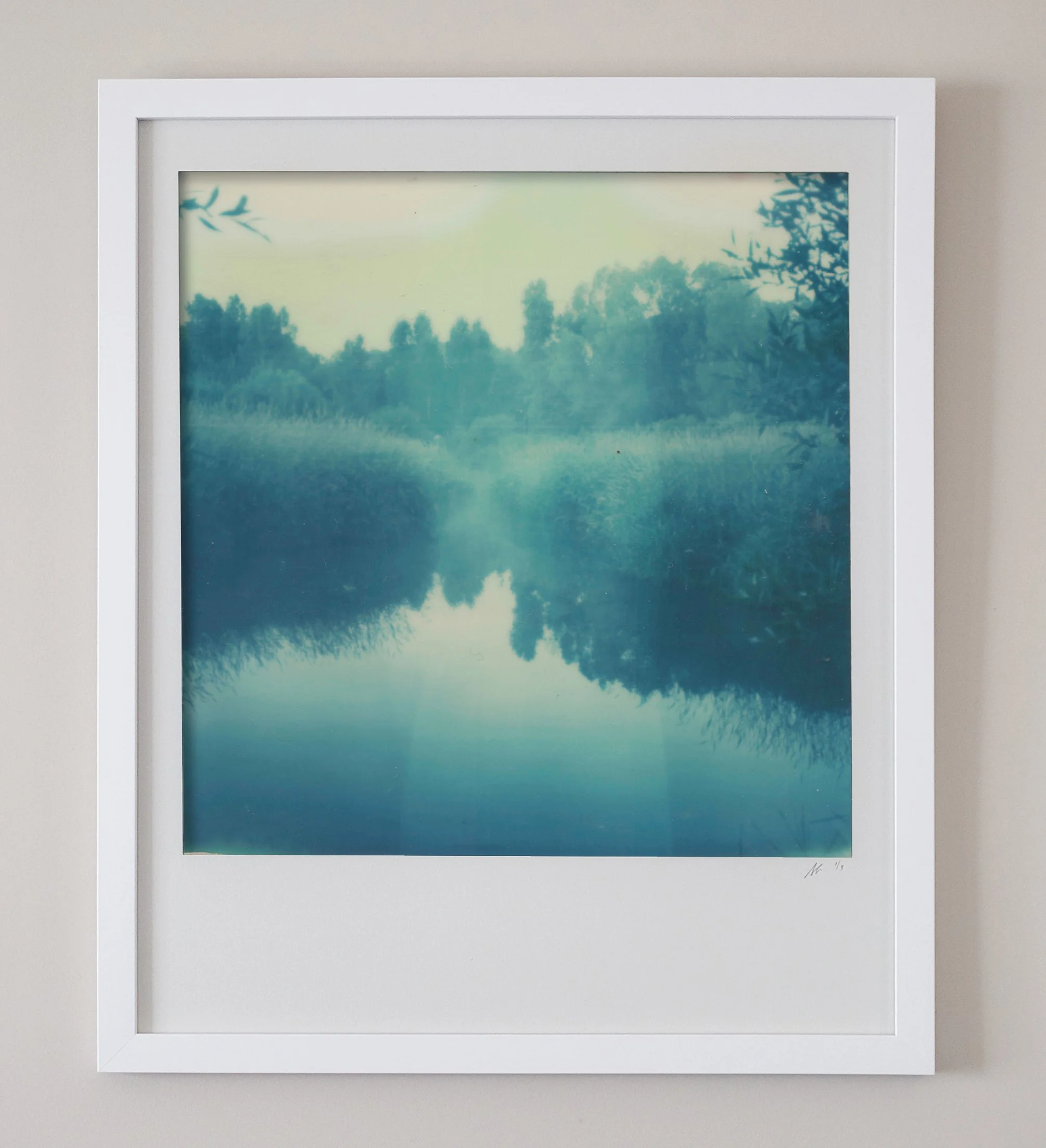 A framed photograph of a calm river or lake surrounded by trees, with the water reflecting the sky and foliage.