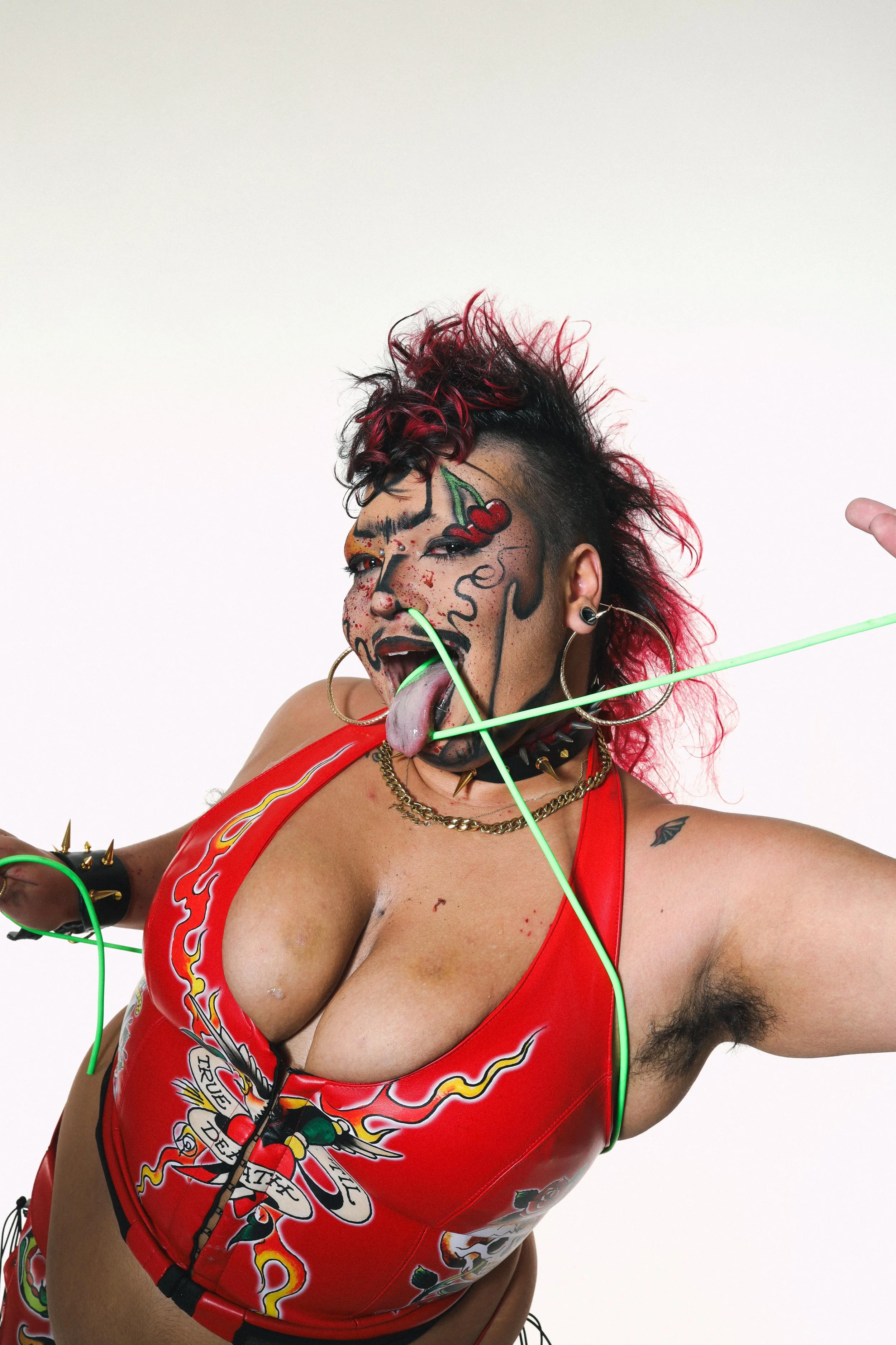 Angel is a fat brown drag king with heavy cartoonish makeup and black and red hair teased up. He is wearing a red faux leather halter top covered in tattoo designs. He has a green tube going in his nose and out of his mouth. 
