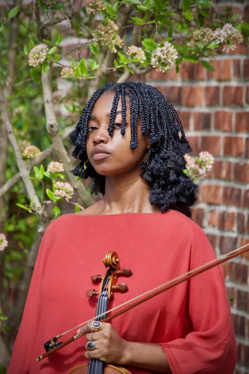 Javen Lara is pictured with her viola in a burnt orange dress shirt, against a brick wall background with flowers ornamented.