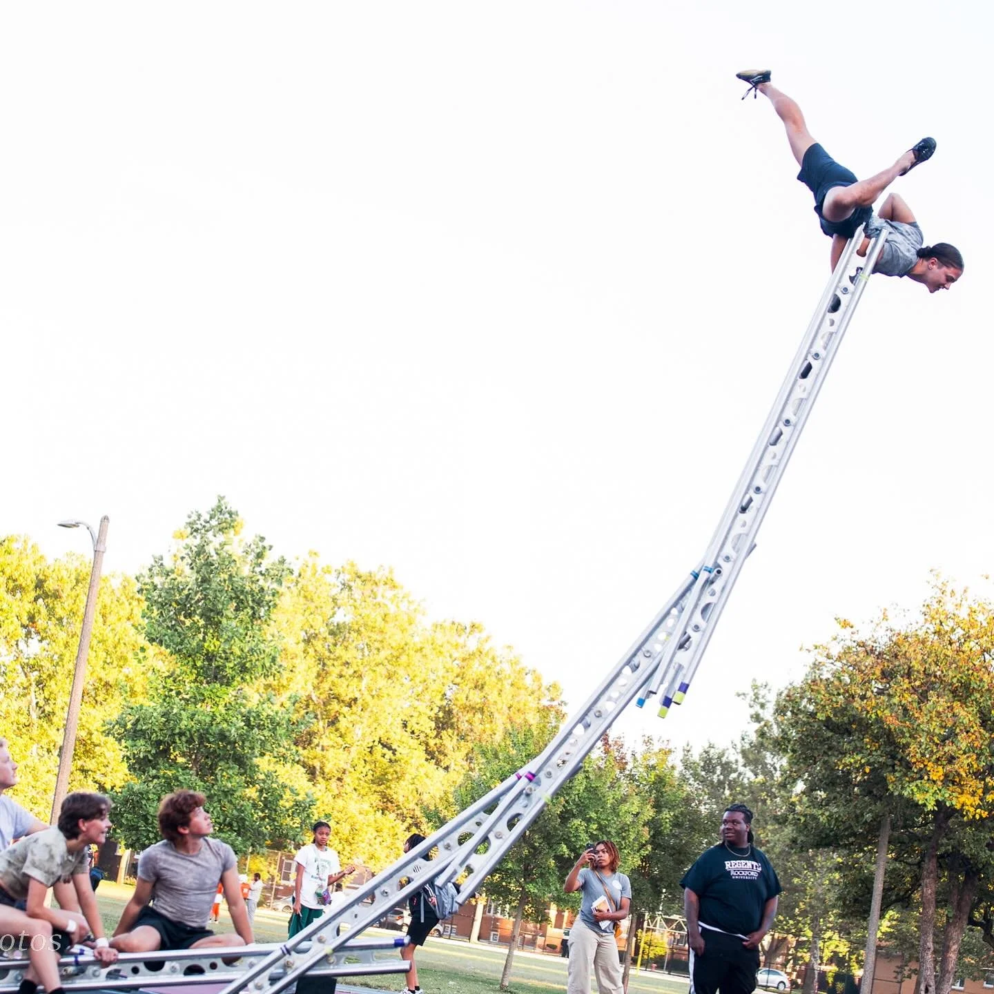 Acrobat does croc pose (partial handstand) on tall ladder sculpture in a park