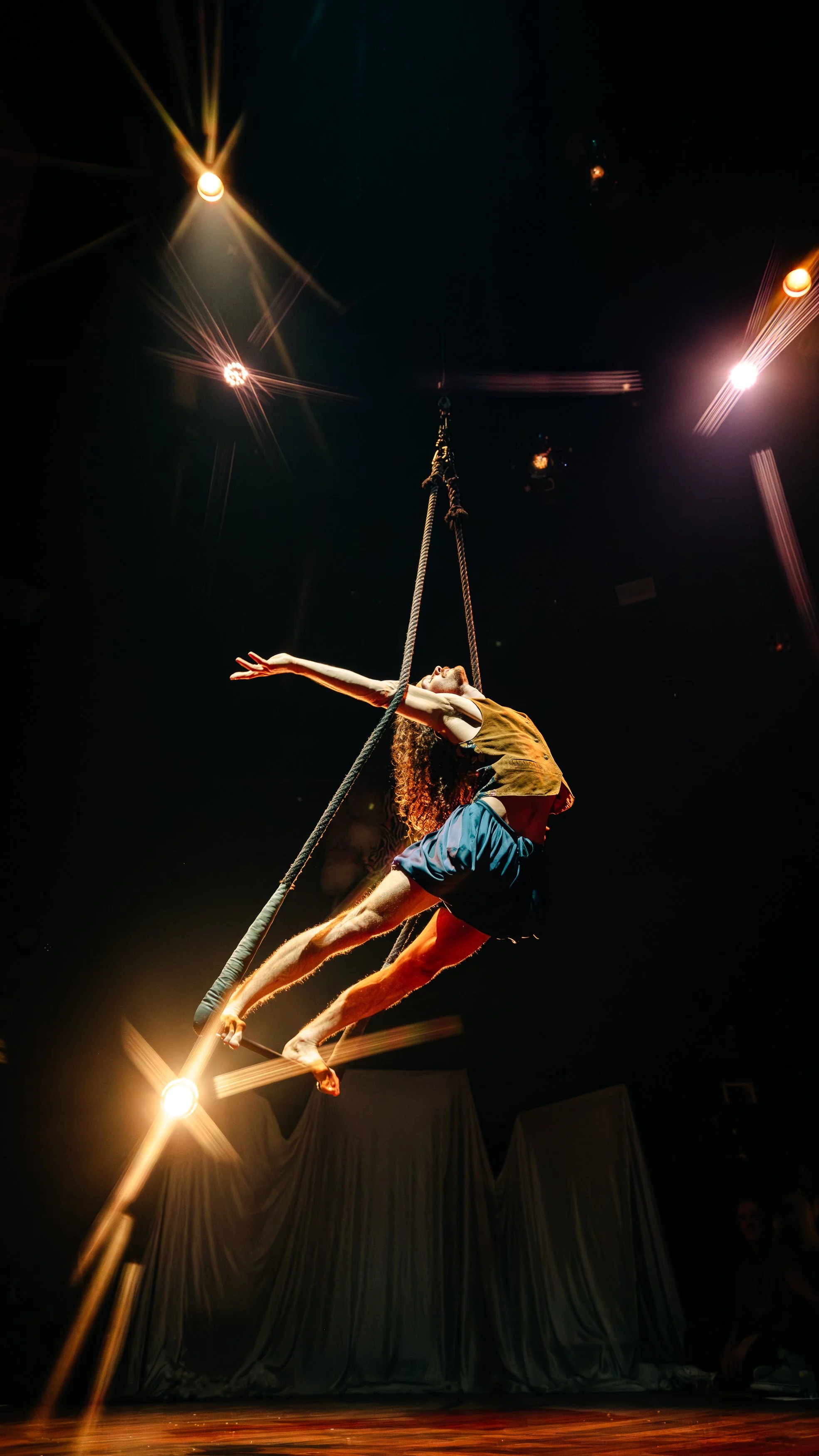 Artist Troy Lingelbach stands on a trapeze with arms outstretched, leaning forward in an arch and held in place by the tension between their arms and the trapeze ropes.
