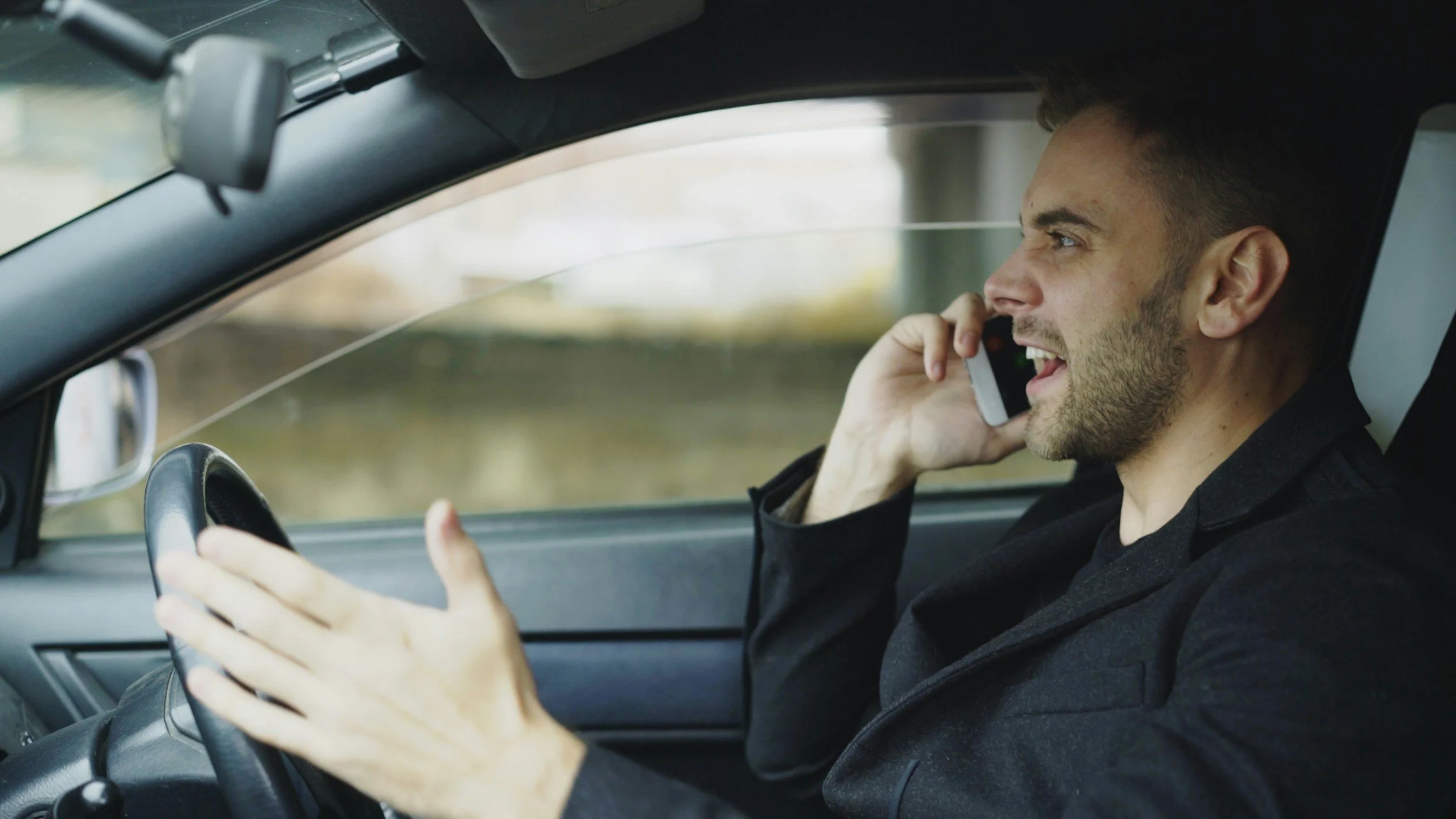 Man driving a car while talking on his phone, gesturing with frustration as if overwhelmed or emotionally reactive.