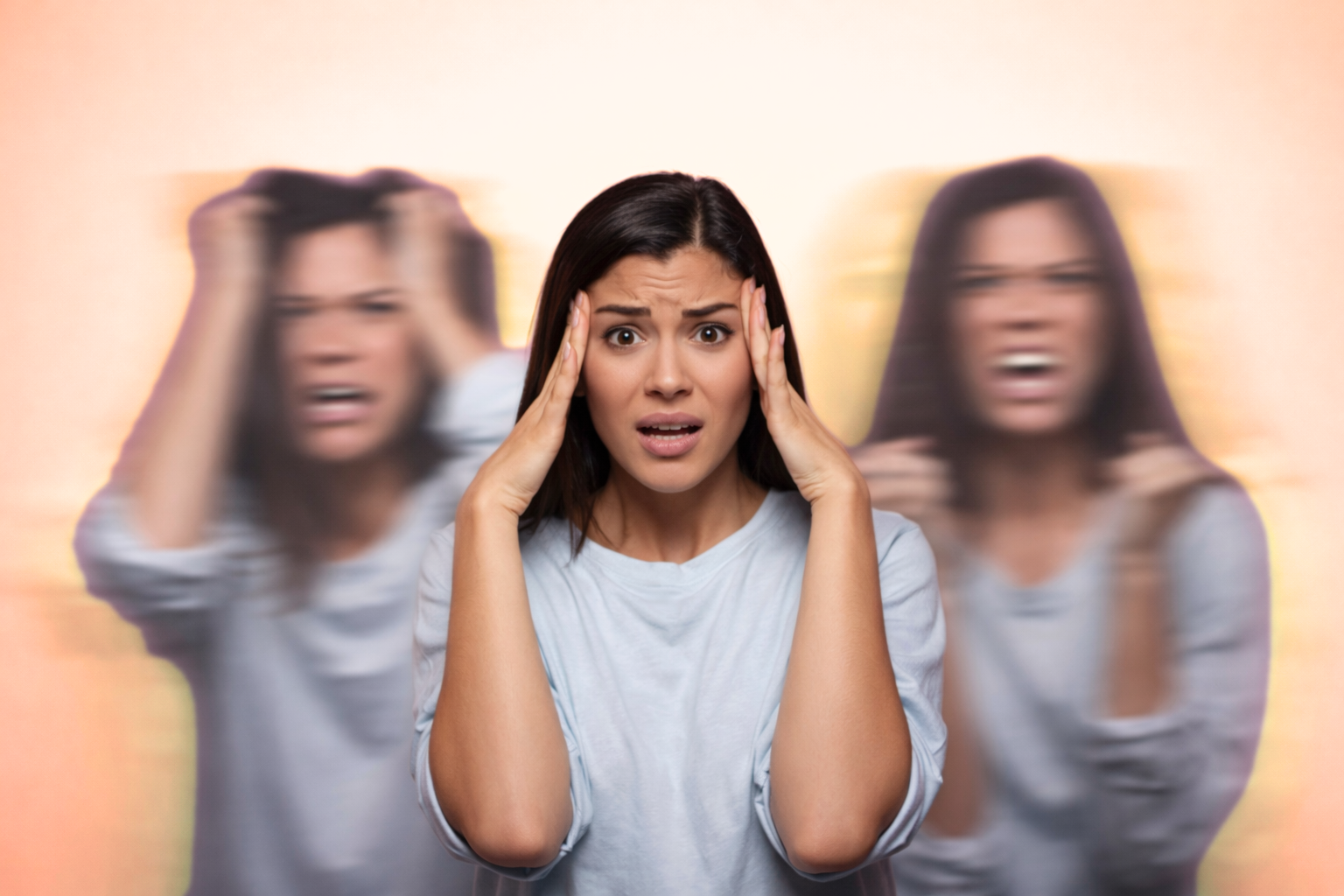 Woman holding her temples with blurred versions of herself on each side, illustrating dissociation and feeling mentally overwhelmed.