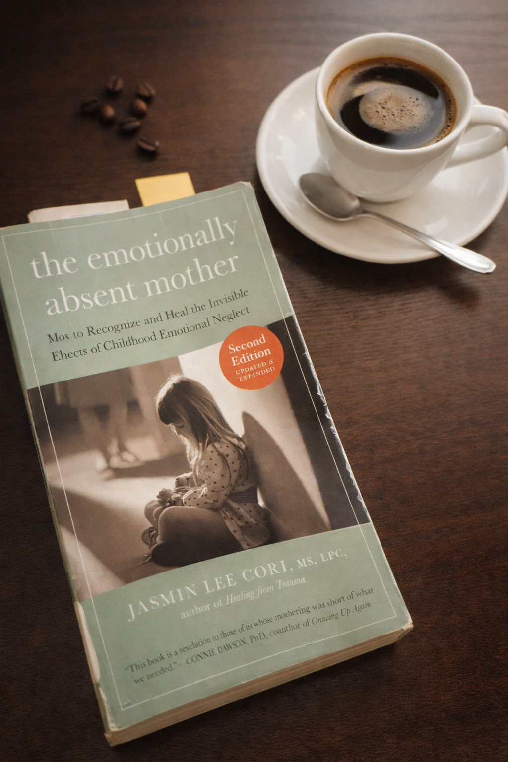 A copy of the book “The Emotionally Absent Mother” by Jasmin Lee Cori on a wooden table next to a cup of coffee, symbolizing reflection and emotional healing.