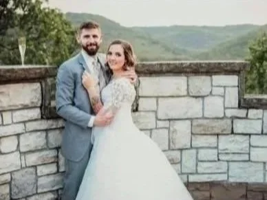 A bride and groom in wedding attire standing outdoors in front of a stone wall, with scenery of hills and trees in the background.