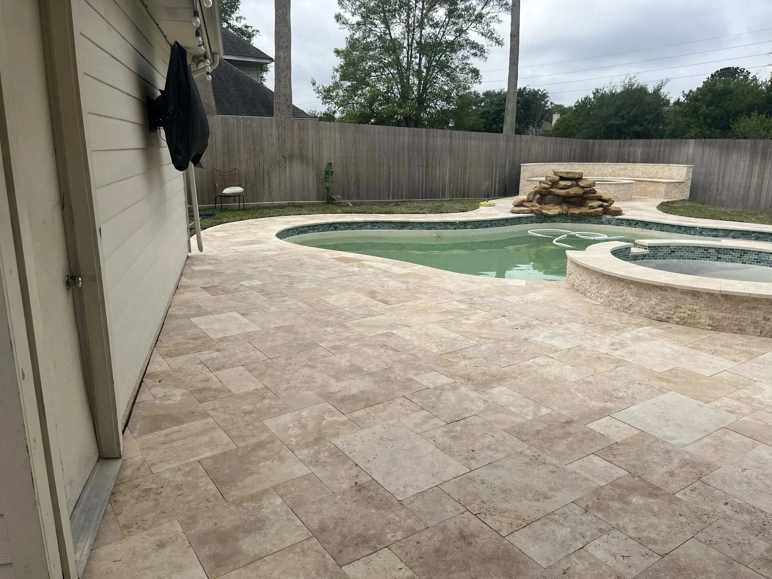 A backyard with a pool, rock waterfall, and a curved seating area built into the fence. The patio is made of beige tiles, and there is a small chair with a white cushion near the house wall. The sky is overcast, and trees and utility poles are visibl