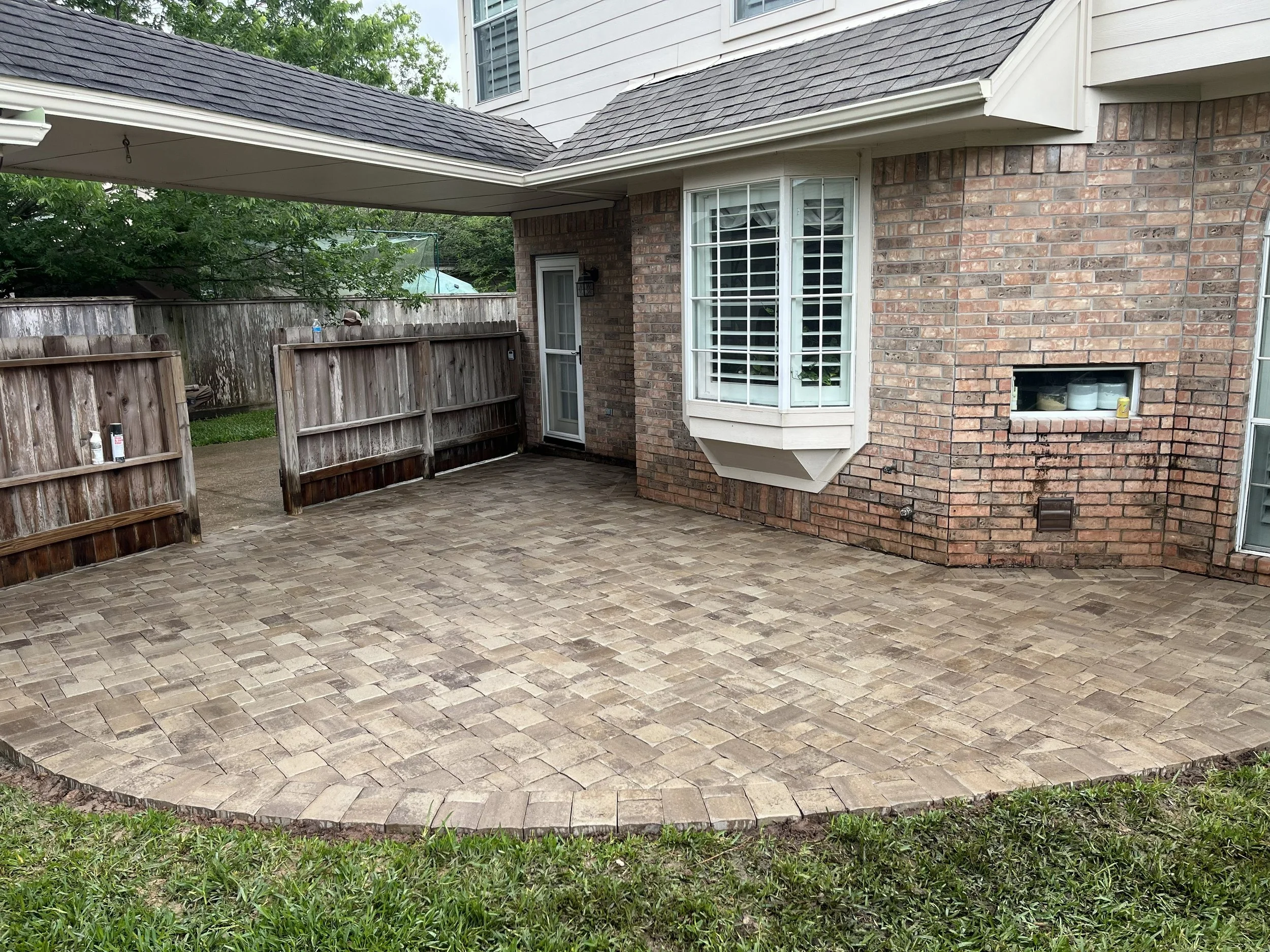 Backyard patio with brick pavers, surrounded by a wooden fence, next to a brick house with white trim and windows, and a small patio door.