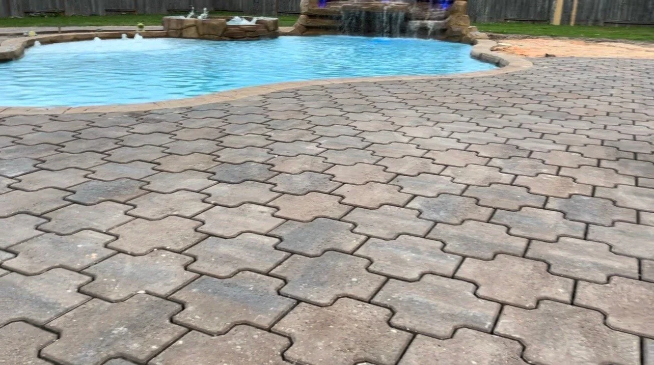 A backyard with a swimming pool and a brick paver patio surrounding it, with a small water feature or waterfall in the background.