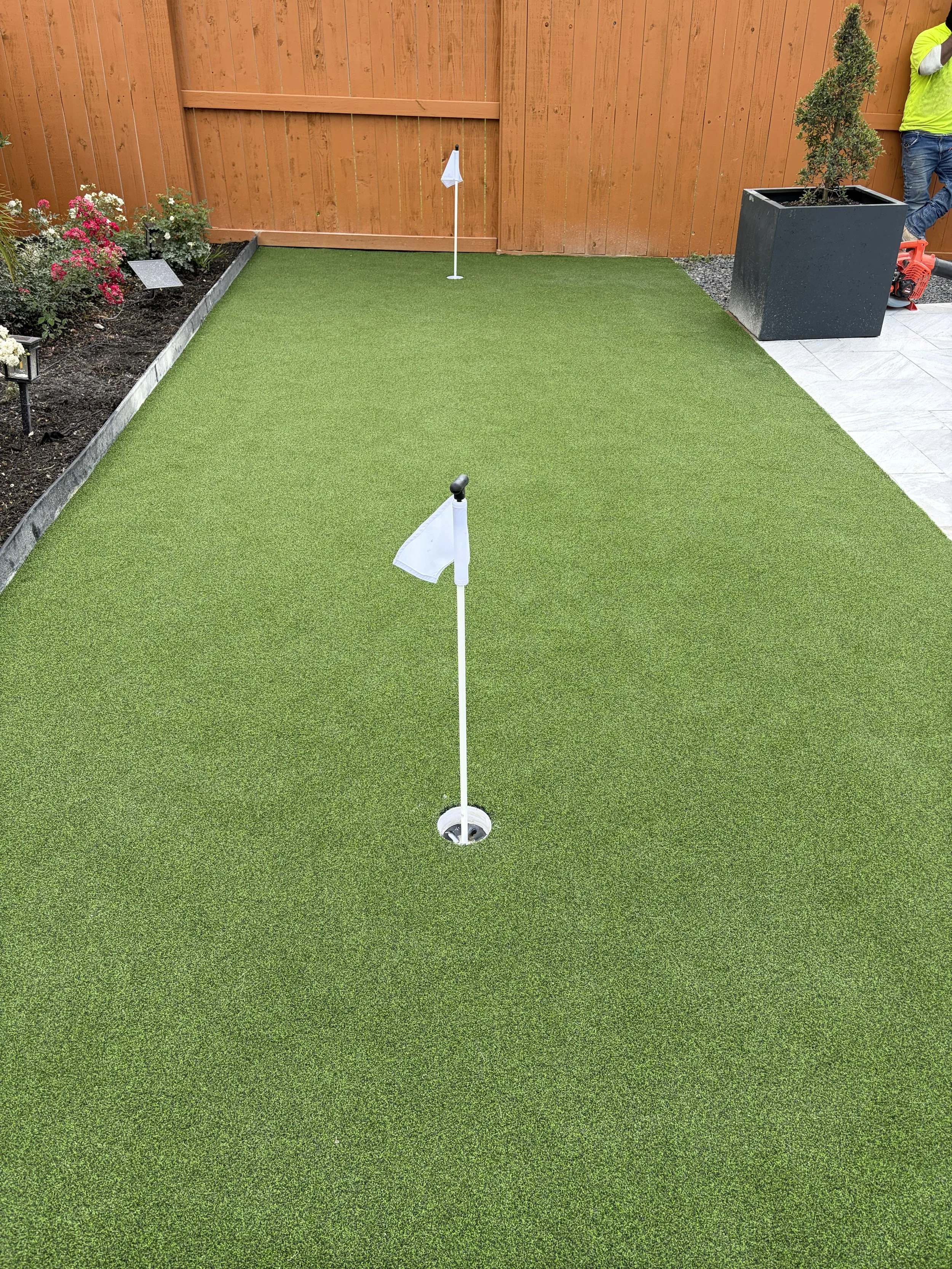 Artificial putting green with two flagsticks, bordered by a flower garden on the left and a concrete patio on the right. A person in a bright yellow shirt is visible in the upper right corner.