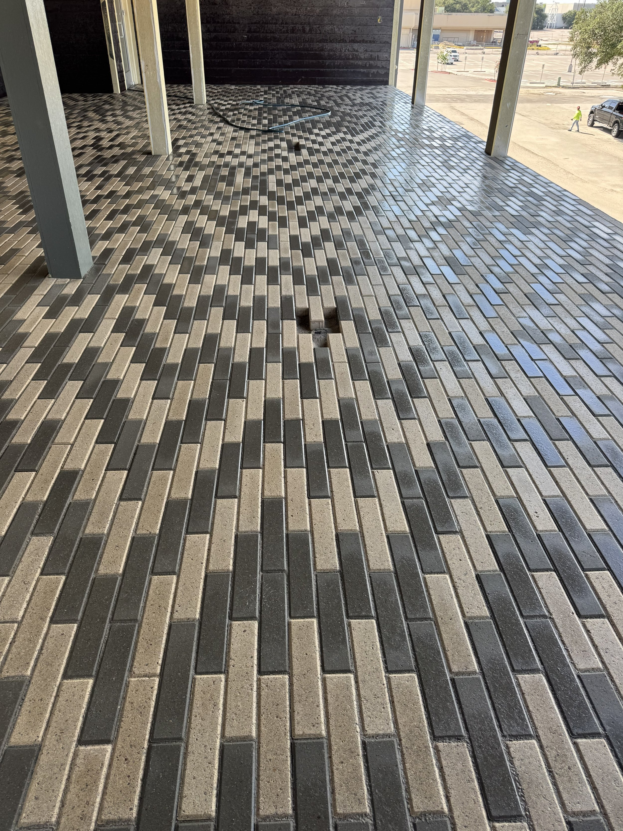 A construction site with a newly installed brick floor and metal posts supporting the structure. In the background, there are parking spaces and a few people walking.