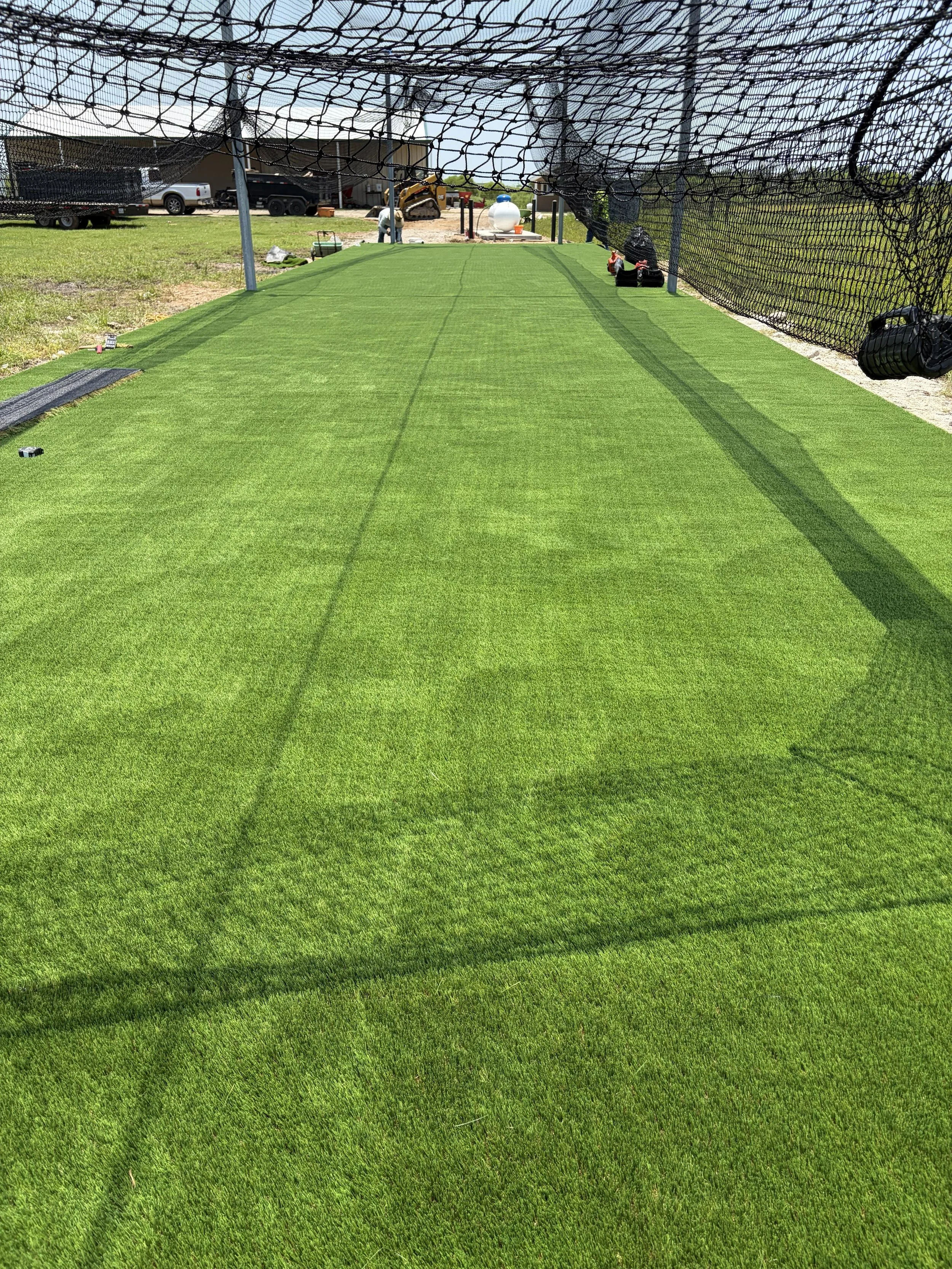A golf practice net with artificial turf and a construction site with a small excavator and construction workers in the background.