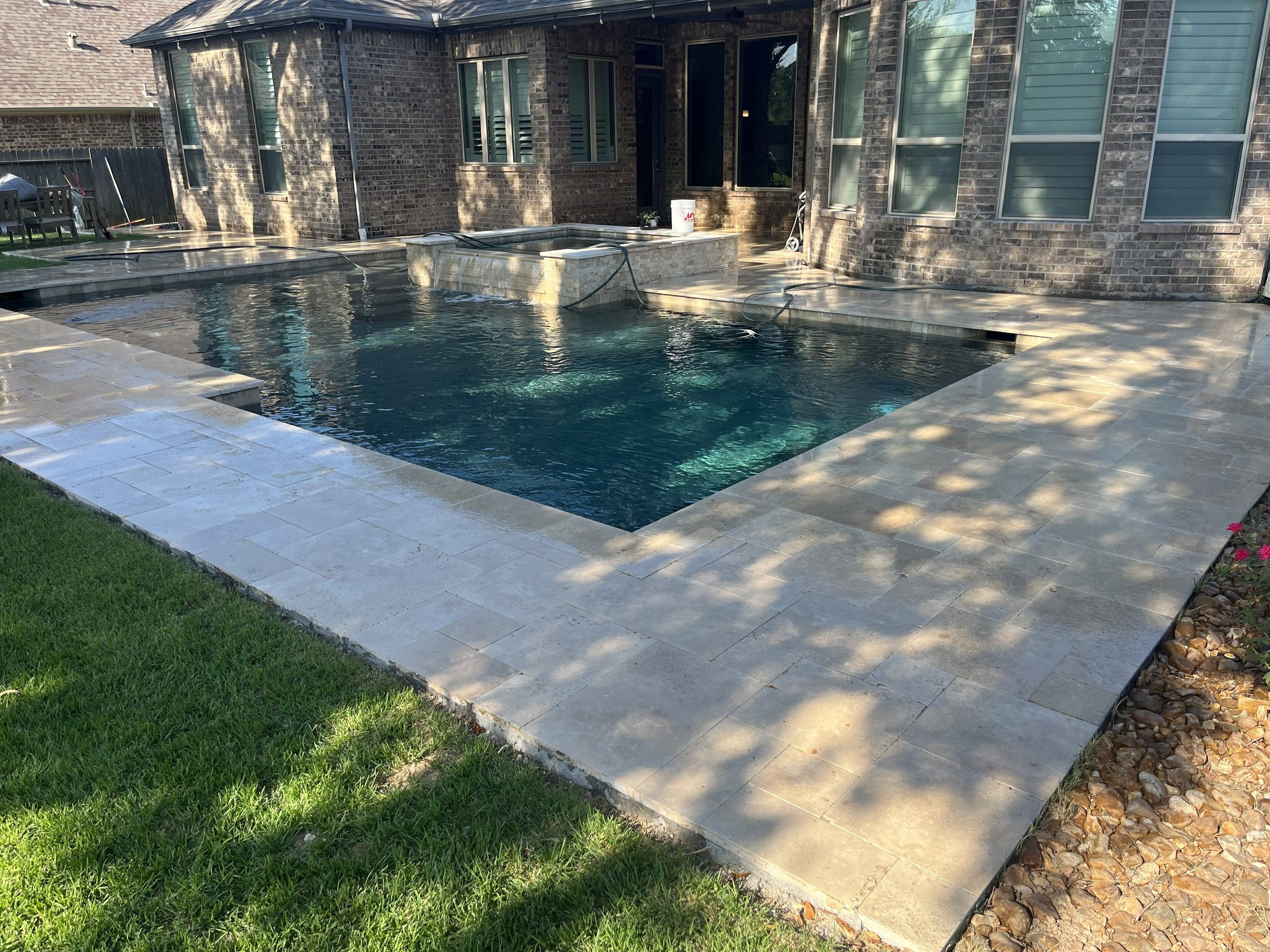 Backyard with a swimming pool, section of patio, brick house with windows and sliding glass door, lawn, and a small garden with flowers.