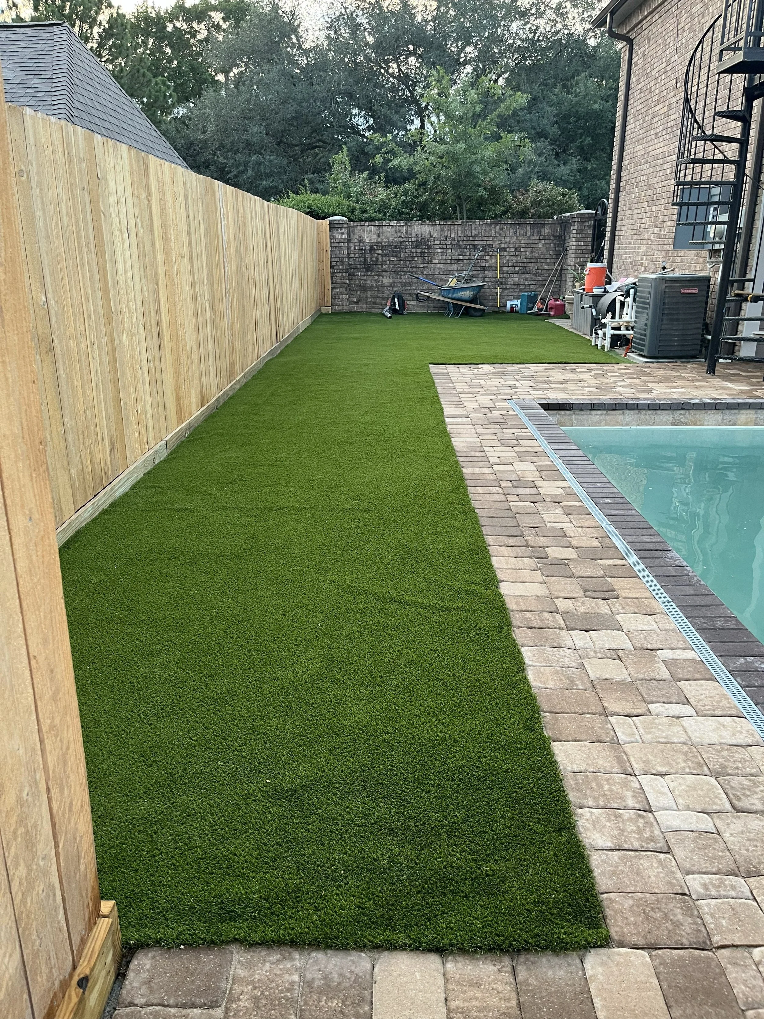 Backyard with artificial grass, brick patio, wooden fence on the left, brick wall in the background, pool on the right, and various gardening tools and equipment near the wall.