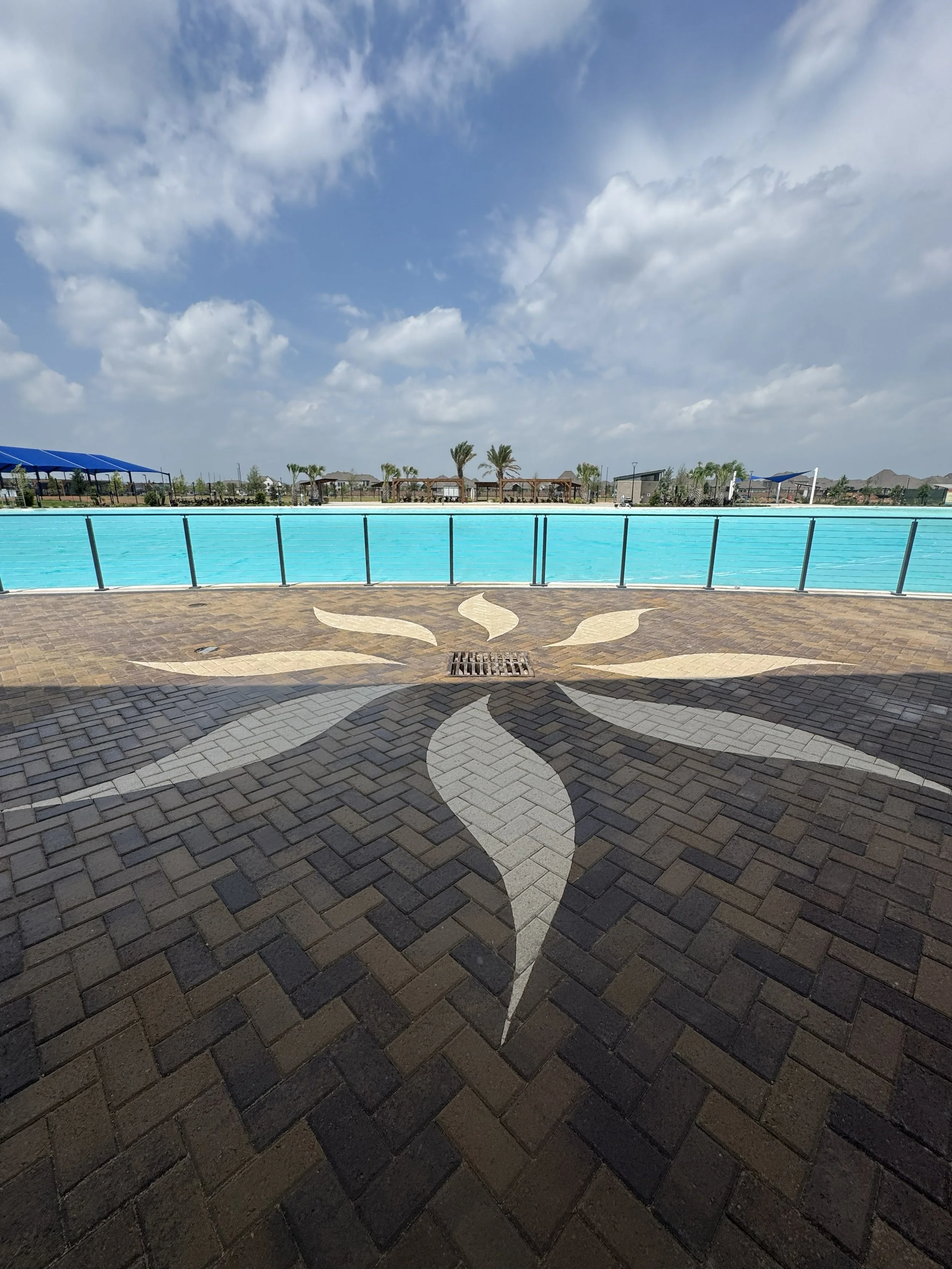 View of a swimming pool with a decorative star-shaped tile pattern on the surrounding brick paved area, under a partly cloudy sky with palm trees and structures in the background.