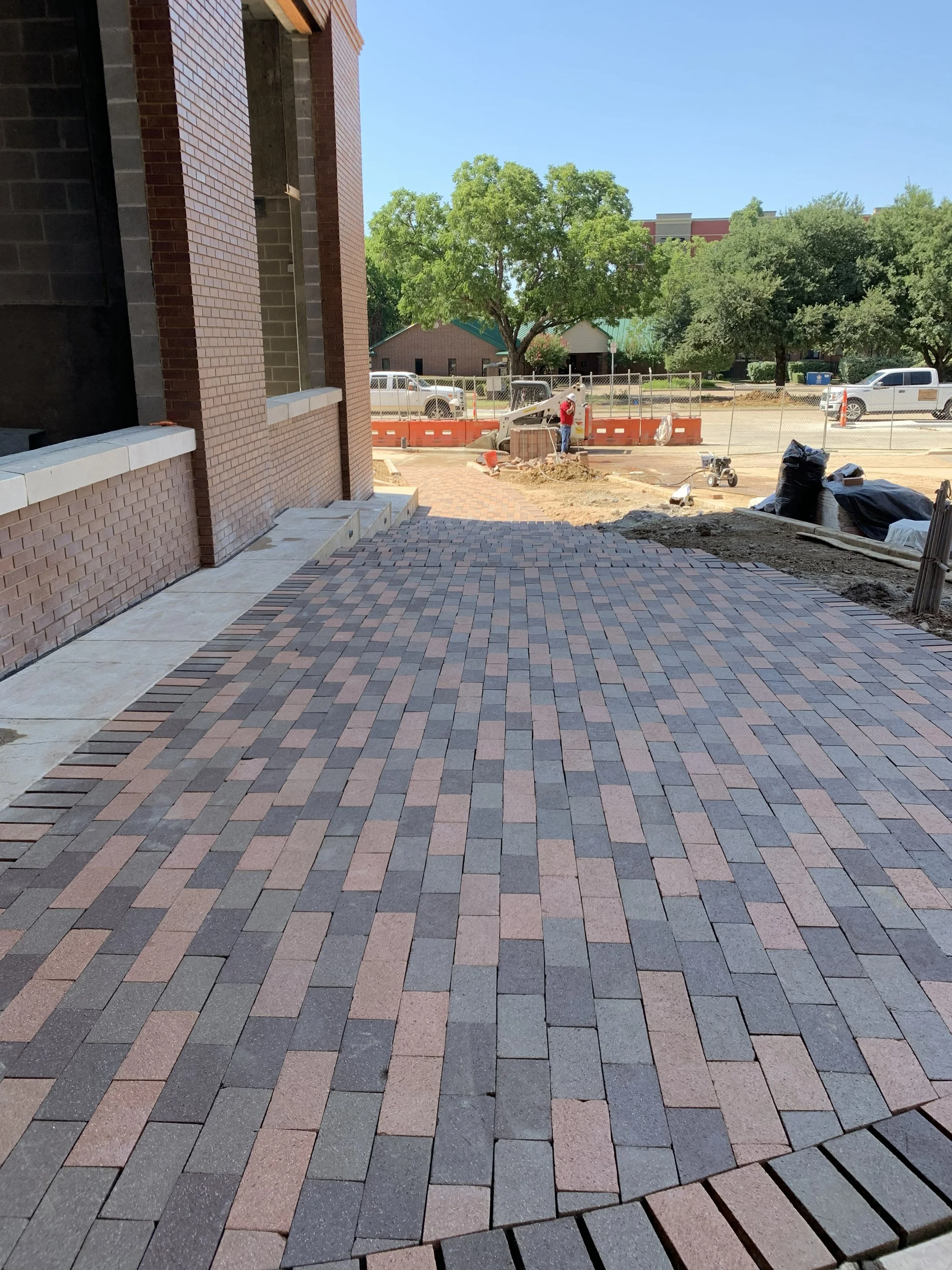 Front view of a construction site with brick pavement, construction materials, and workers, with trees and parked vehicles in the background under a clear blue sky.