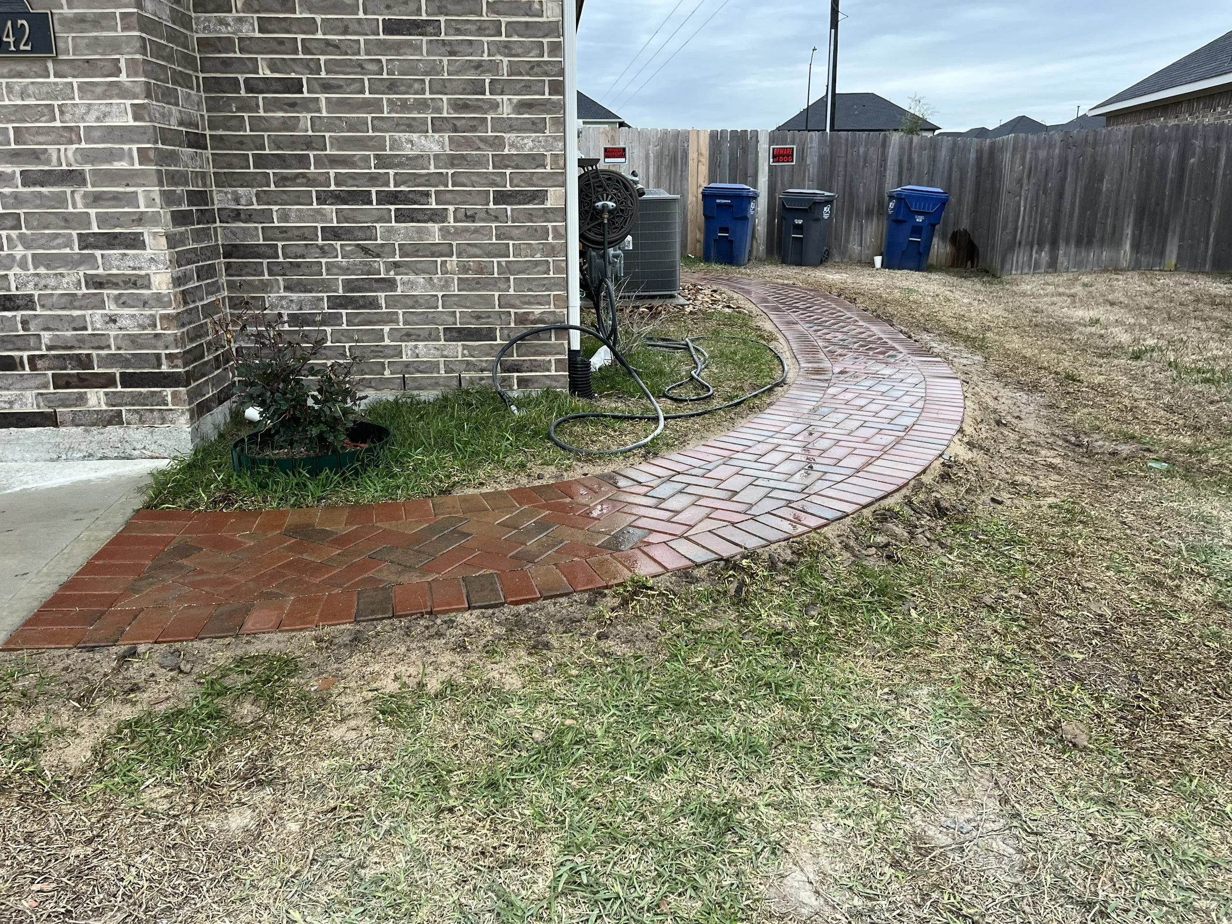 Backyard with brick pathway, trash bins, gardening hose, and a wooden fence. Part of a house with brick siding is visible on the left.