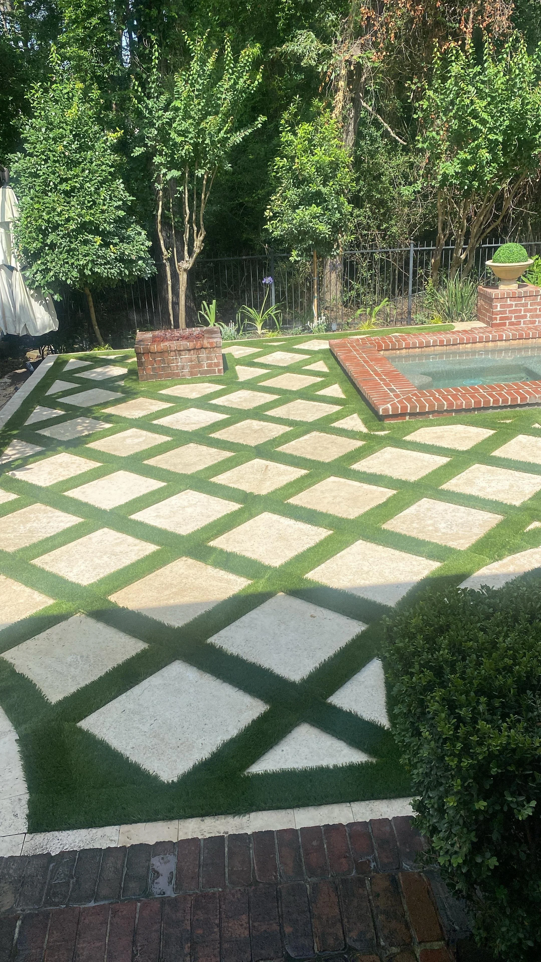 A backyard patio with a patterned concrete and grass design, trees, and a small pool with a brick border. There are also decorative plants and brick garden borders.