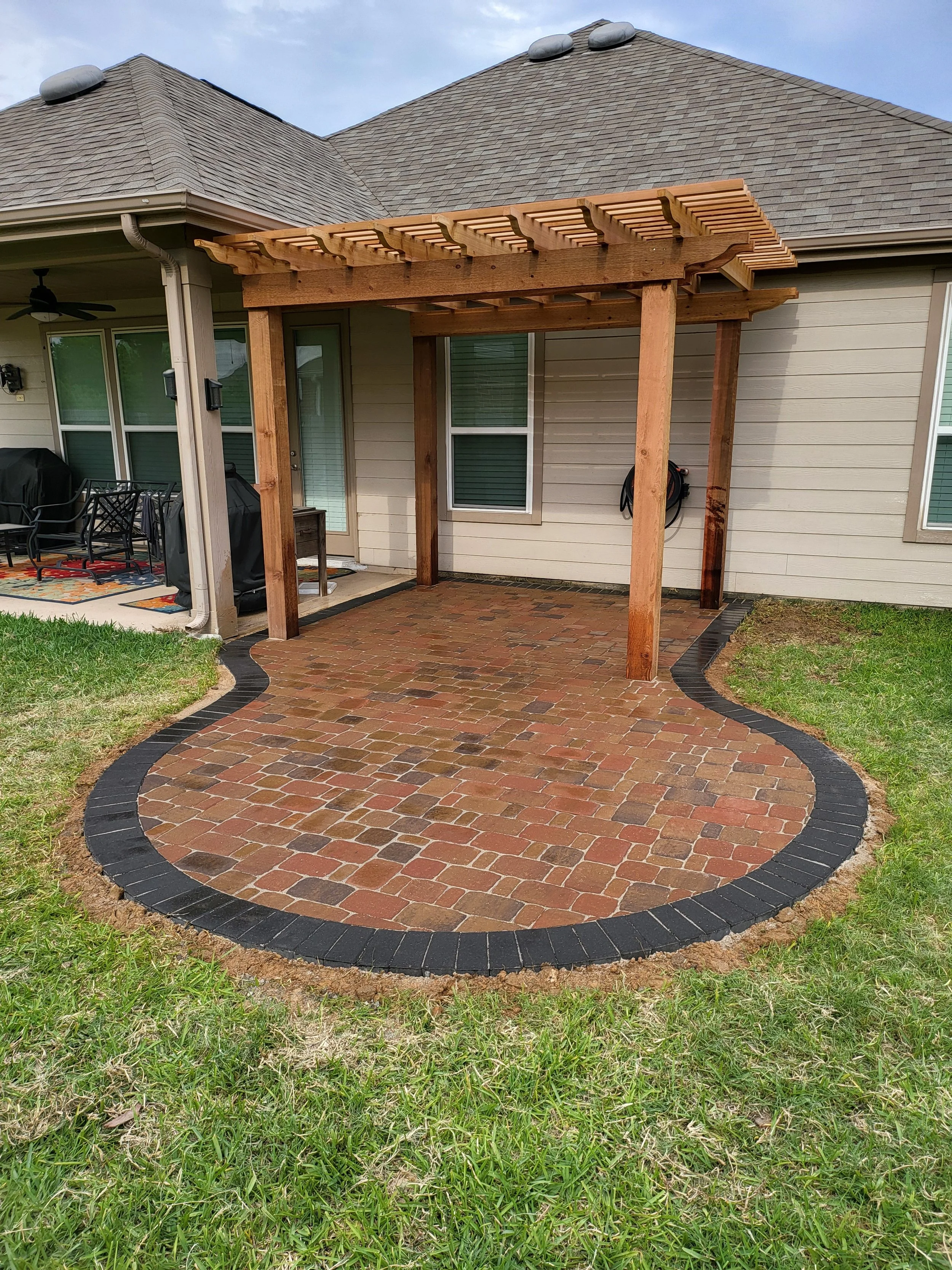Backyard patio with a brick and paver pathway, partially covered by a wooden pergola, with a house in the background featuring sliding glass doors, windows, and a beige exterior with a shingled roof.