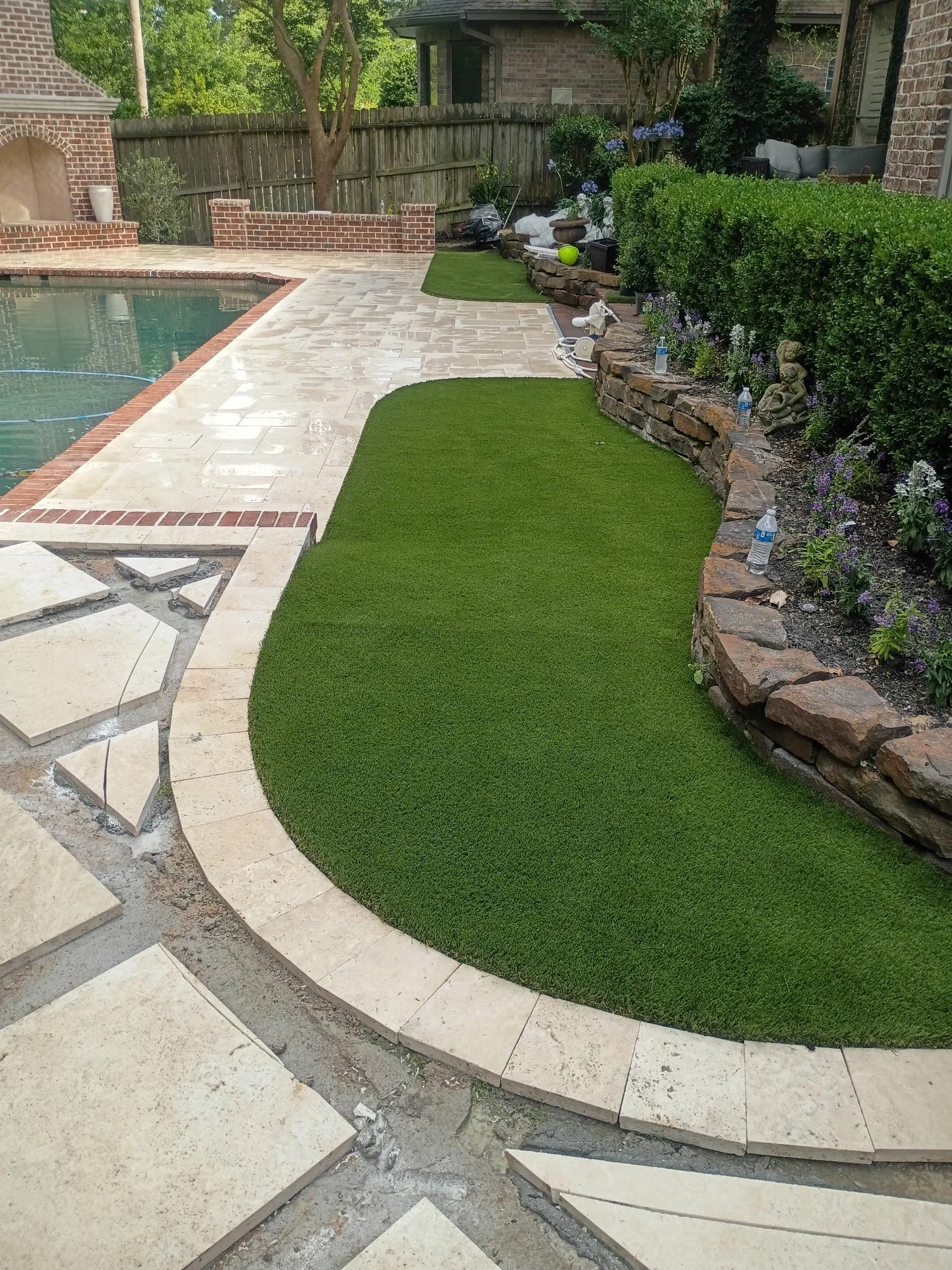 Backyard with a swimming pool, brick patio, artificial grass area, stone retaining wall with plants, and outdoor furniture.